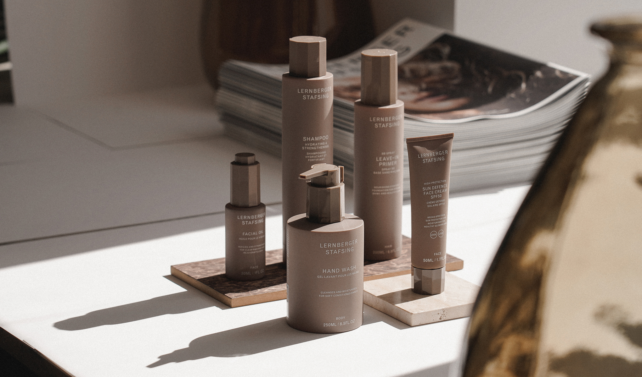 Useita Lernberger Stafsing -tuotteita, kuten shampoo, käsienpesugeeli, kasvoöljy ja aurinkosuojavoide, seisovat pöydällä. Taustalla on pino lehtiä.