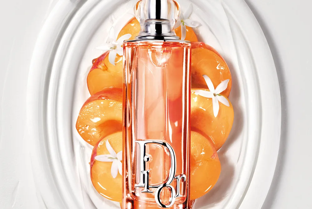 Dior Addict Lip Glow Oil i orange flaska placerad på ett dekorativt vitt fat omgivet av aprikos och små vita blommor.