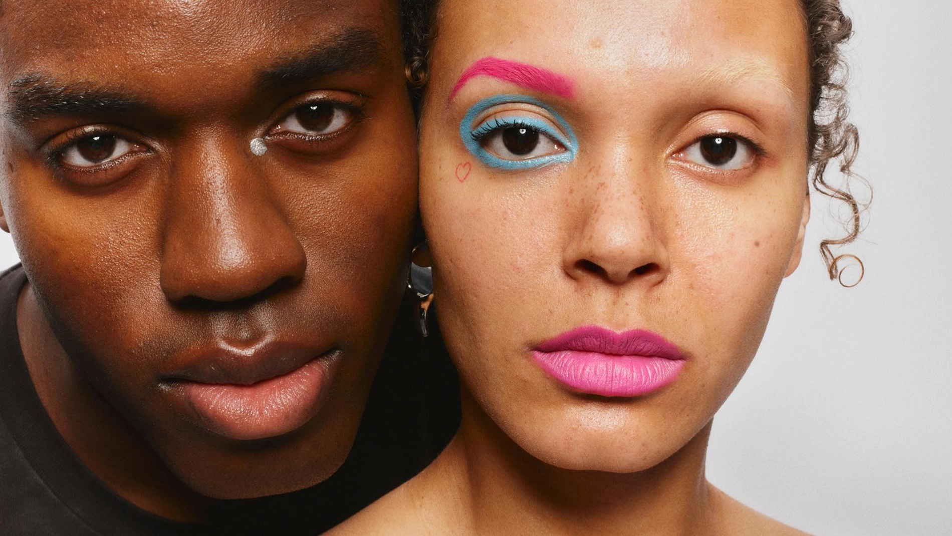 Två personer poserar nära varandra; en har färgstark och kreativ ögonmakeup i rosa och blått.