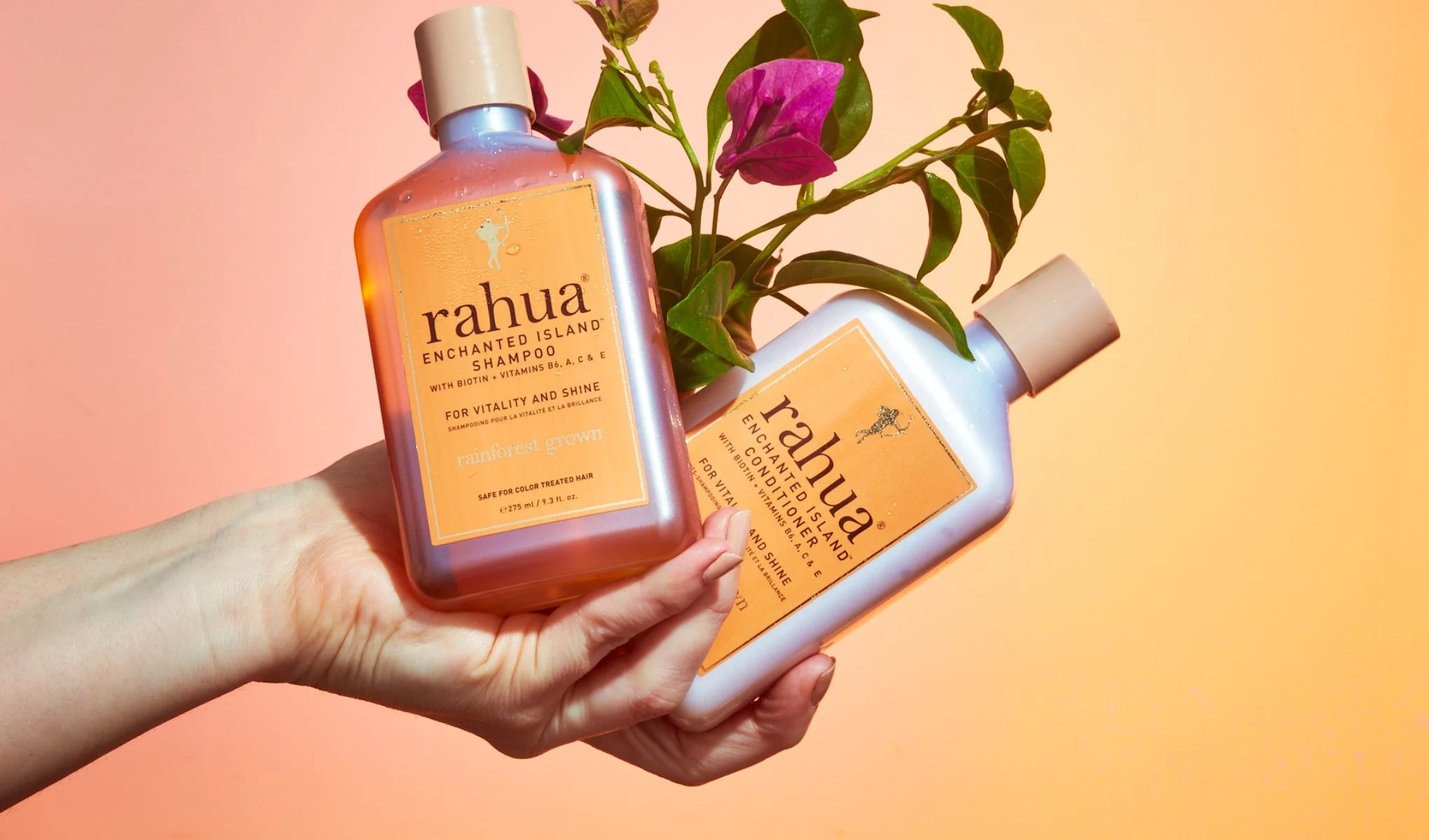 En hand som håller två flaskor av Rahuas Enchanted Island Shampoo och Conditioner. Flaskorna har orange etiketter och är dekorerade med en blommande växt. Bakgrunden är en gradient av rosa och orange.