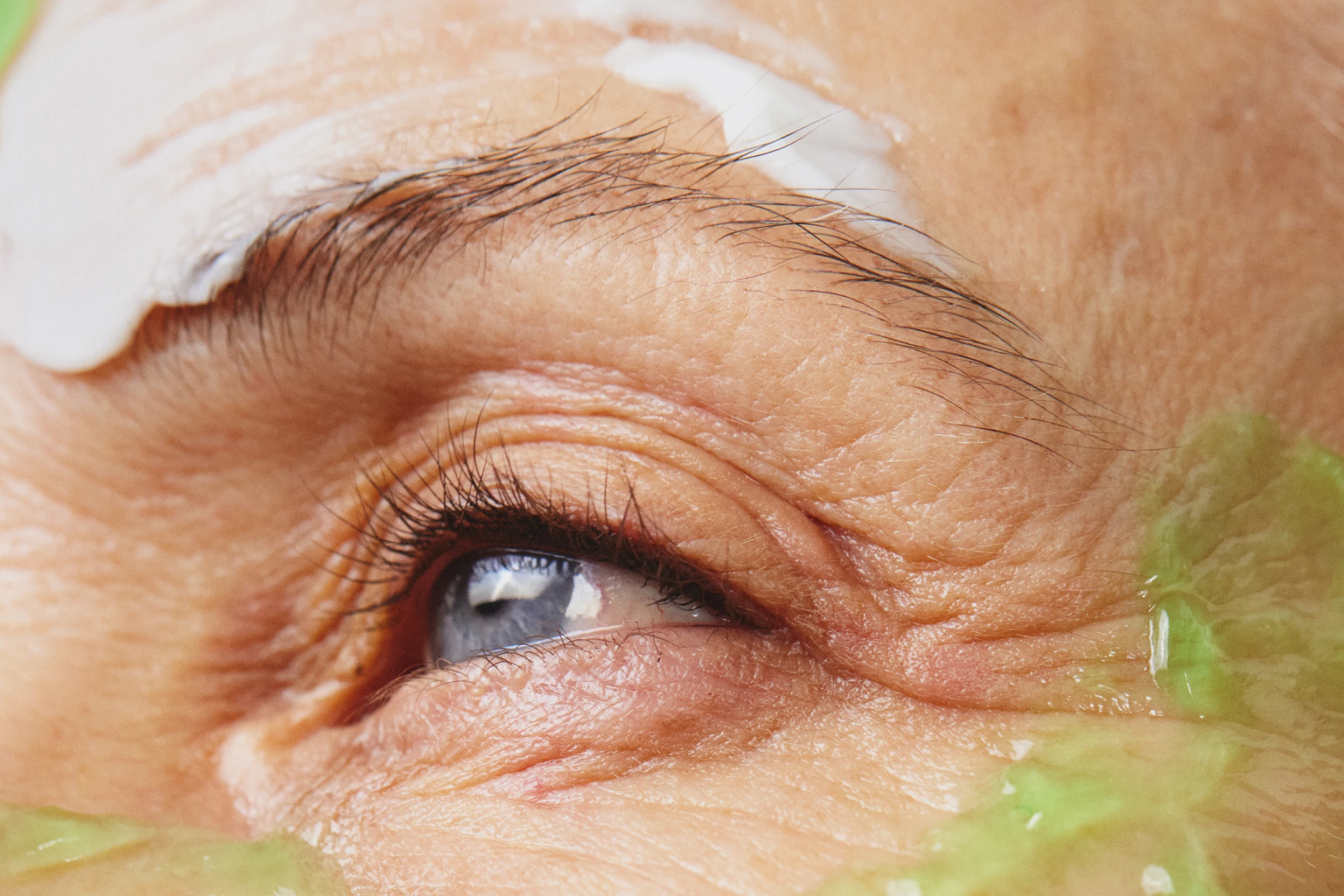 Lähikuva silmästä ja silmänympärysihosta, jossa vihreää geelipohjaista kasvonaamiota silmän ympärillä, näkyviä ihopoimuja ja luonnolliset ripset vaaleaa taustaa vasten.
