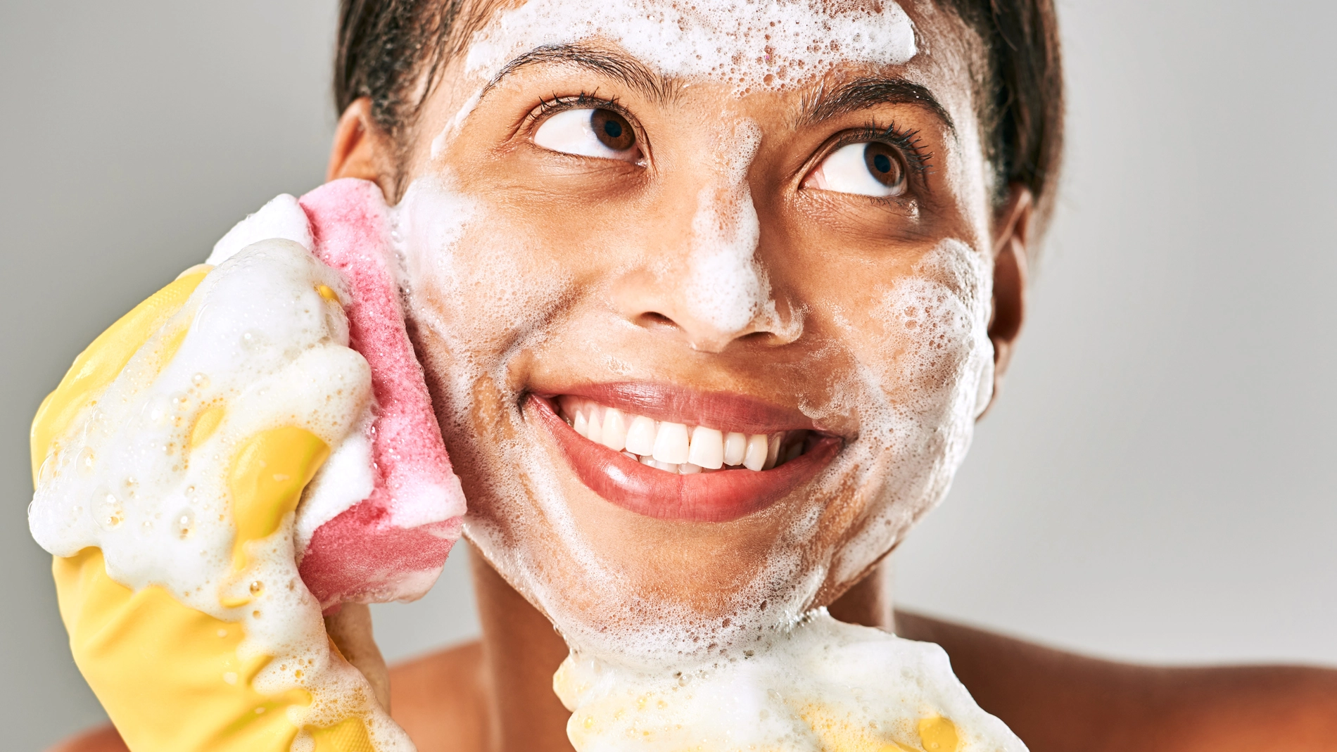 Person med uppsatt hår tvättar ansiktet med rikligt lödder. Hen håller en rosa och gul tvättsvamp mot kinden och bär gula rengöringshandskar. Vitt skum täcker panna, kinder och haka mot ljus bakgrund.