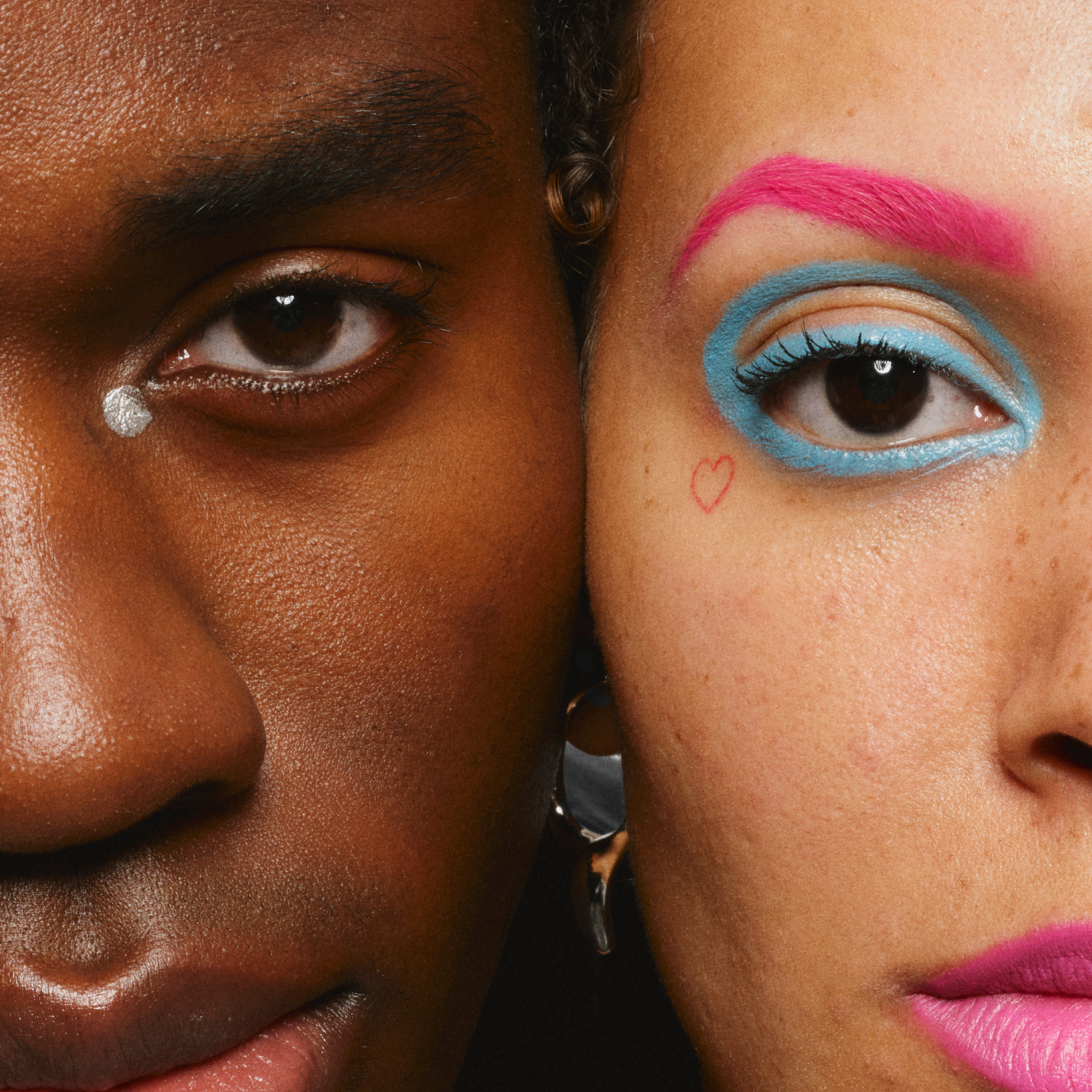 Två personer med kreativ ögonmakeup, en med en silverpunkt under ögat och den andra med blå ögonskugga och rosa ögonbryn samt ett litet hjärta på kinden.