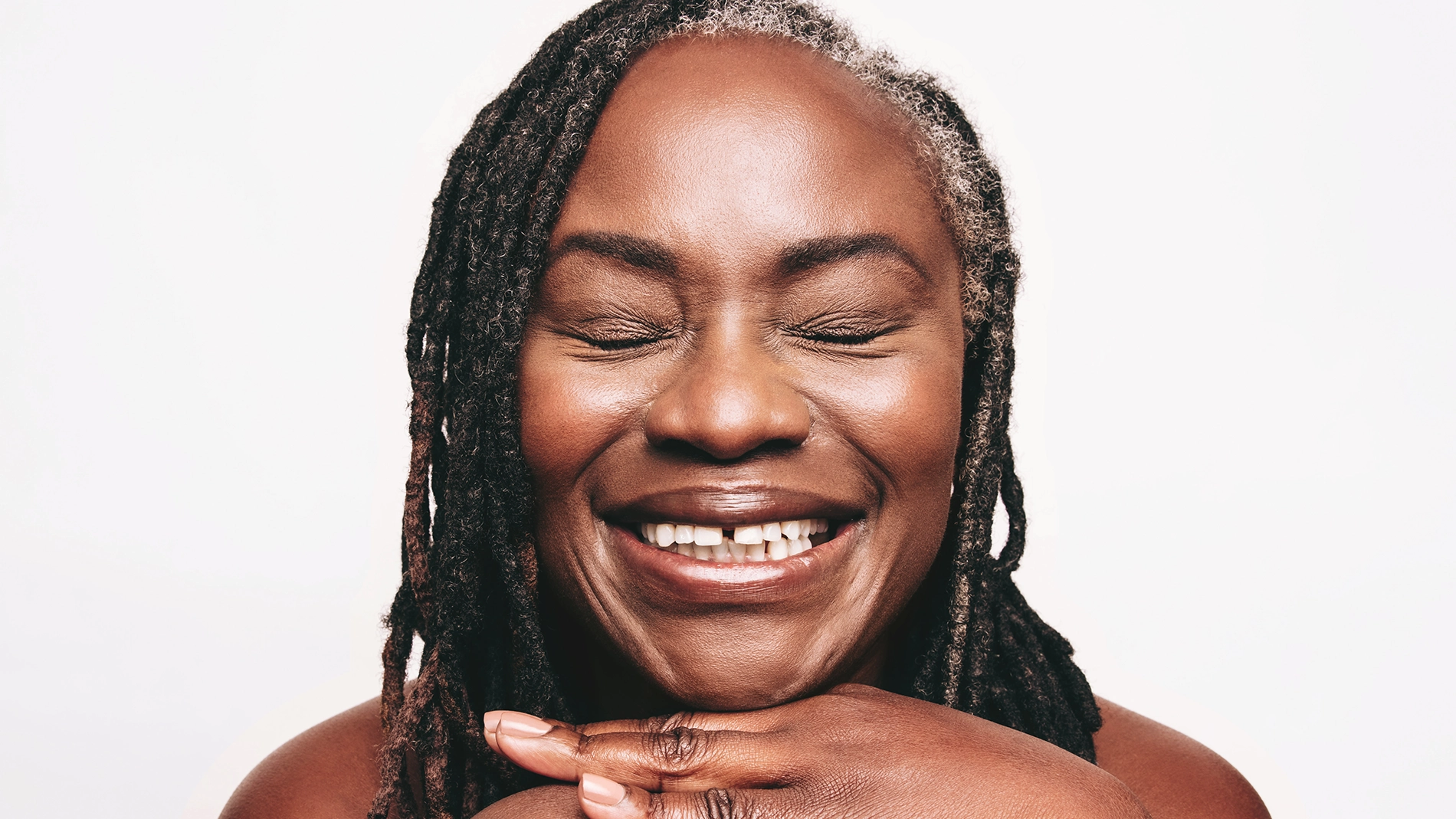 Närbild av person med slutna ögon och naturlig, strålande hud, med fokus på hudvård, lyster och jämn hudfinish. Händerna är placerade under hakan och framhäver ansiktets form.