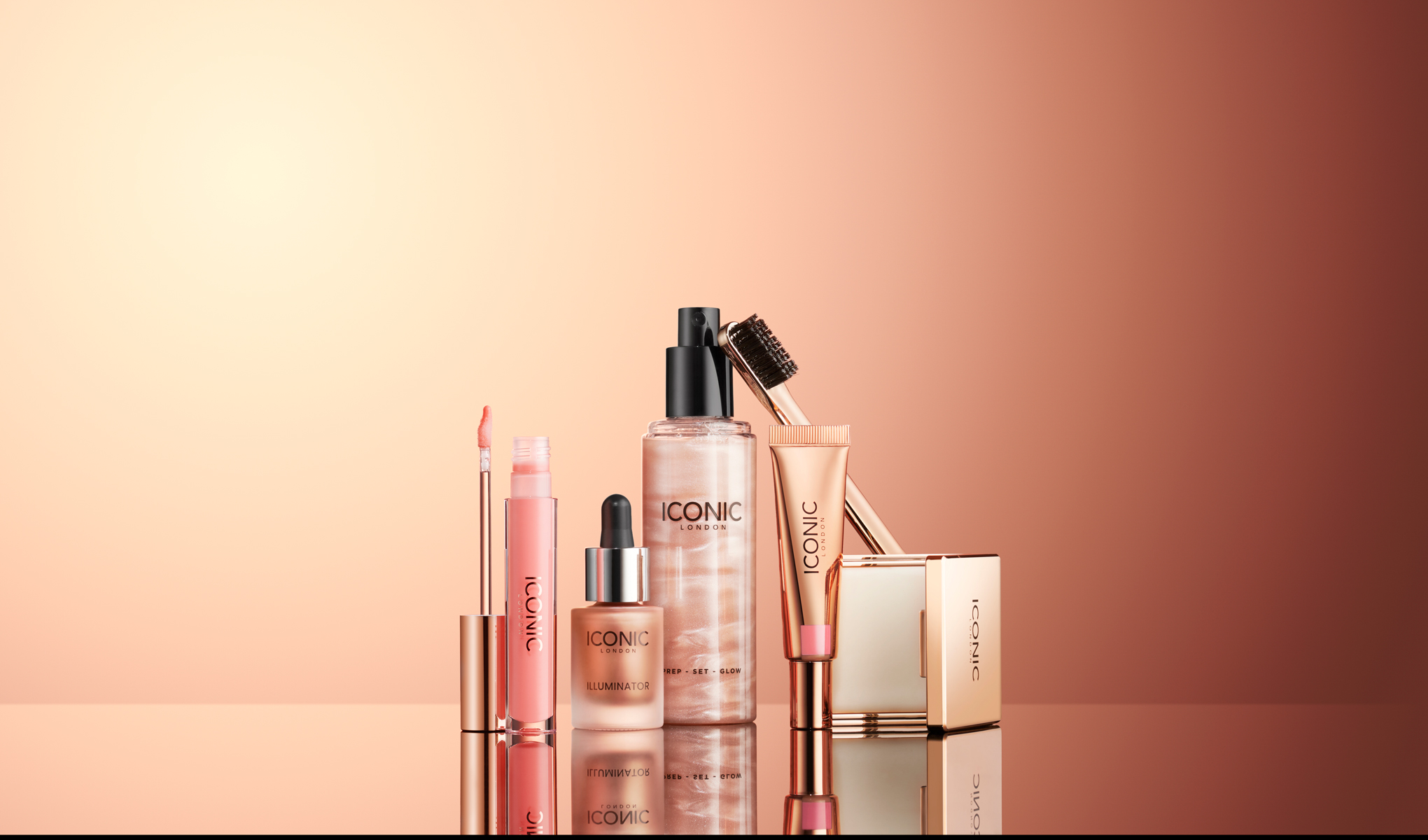 Flera kosmetikaprodukter från Iconic London, inklusive läppglans, highlighter, ansiktsmist, ögonbrynsborste och foundation. Produkterna är arrangerade på en reflekterande yta med en bakgrund i varma, rosa toner.