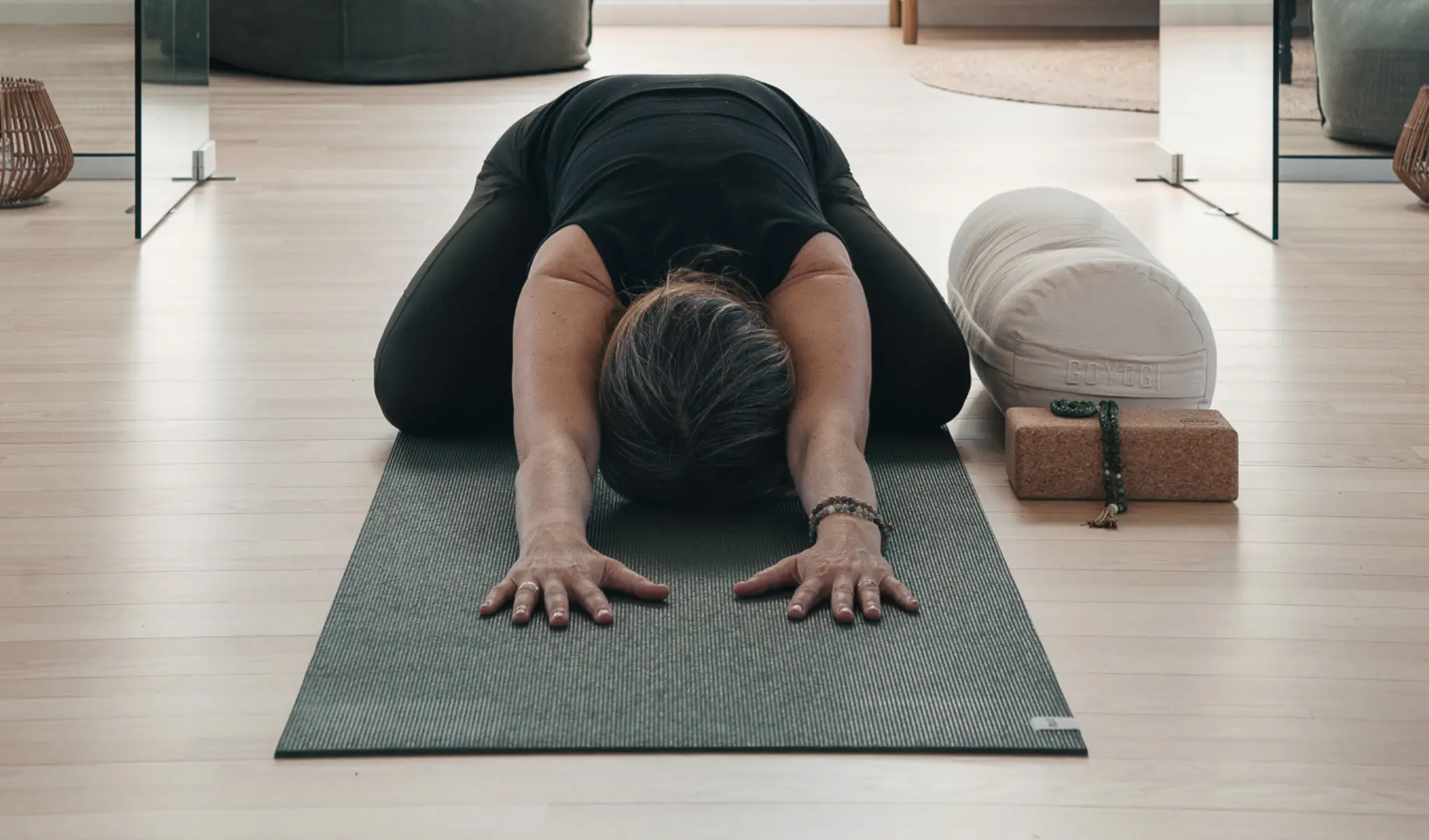 Person i svart träningskläder utför en framåtfällning/yogaposition på en yogamatta i ett ljust rum, med armarna utsträckta framåt och pannan mot mattan. Bredvid ligger en rund yogakudde, ett yogablock och ett radband som förstärker en lugn wellness- och träningsmiljö.