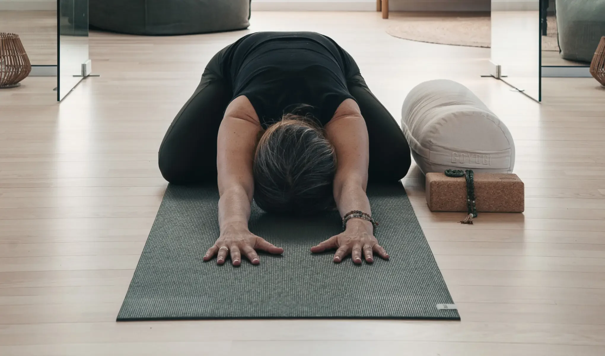 Person i svart träningskläder utför en framåtfällning/yogaposition på en yogamatta i ett ljust rum, med armarna utsträckta framåt och pannan mot mattan. Bredvid ligger en rund yogakudde, ett yogablock och ett radband som förstärker en lugn wellness- och träningsmiljö.