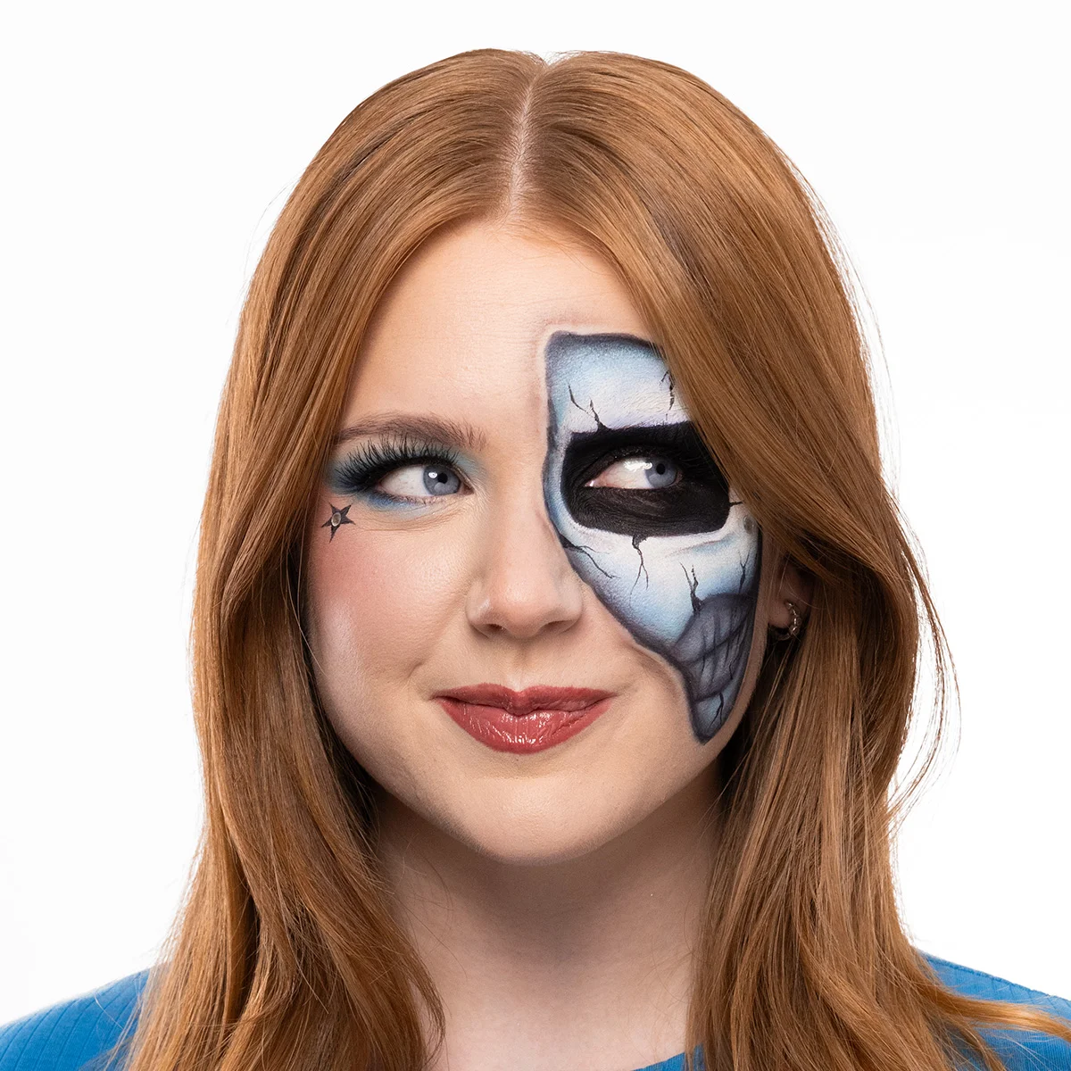 En person med halvskelettinspirerad face paint där ena ansiktshalvan har blå och svarta målade sprickor och ett mörkt ögonhålsmotiv, medan den andra sidan har blå ögonmakeup och en liten stjärndetalj vid ögat.