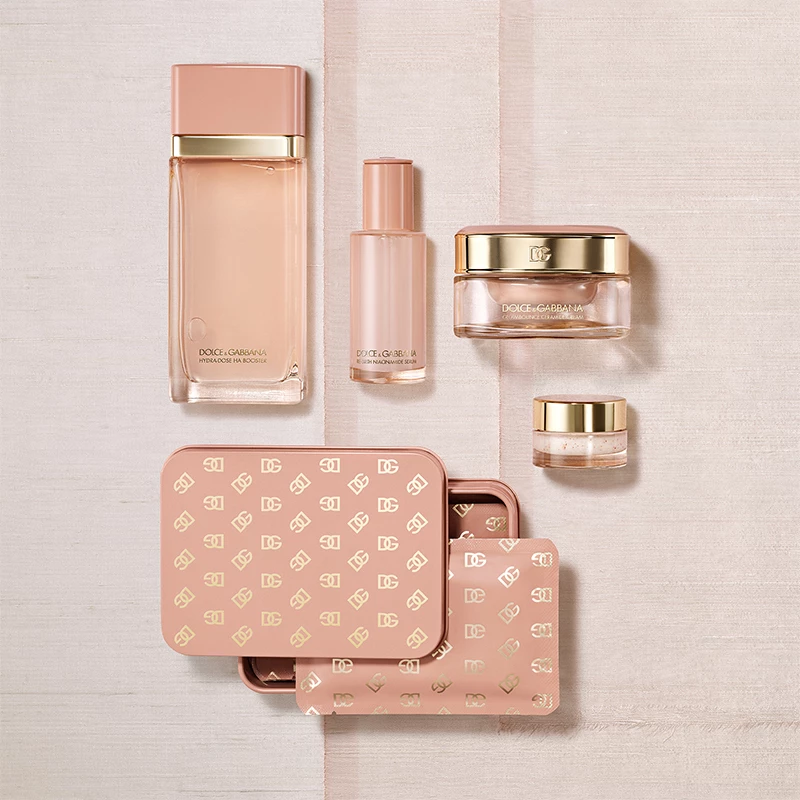 Flera kosmetikprodukter i rosa och guld, inklusive parfymflaskor, krämer och en metallask med Dolce & Gabbana-logotyper, ligger på en ljus yta.