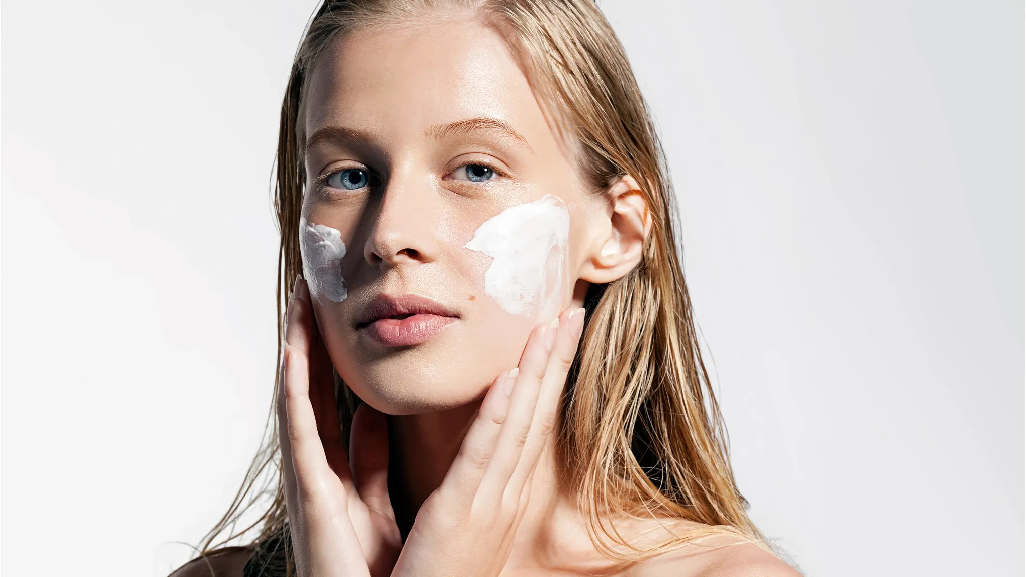 En person med blont hår och händerna vid ansiktet, med vit creme på kinderna