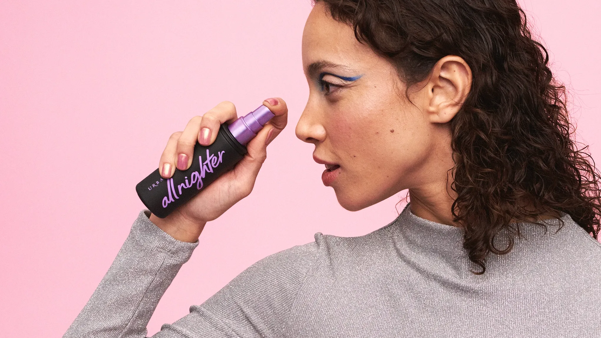 En kvinna med lockigt hår och makeup i profil, som visar upp en All Nighter Setting Spray från Urban Decay, mot en rosa bakgrund.
