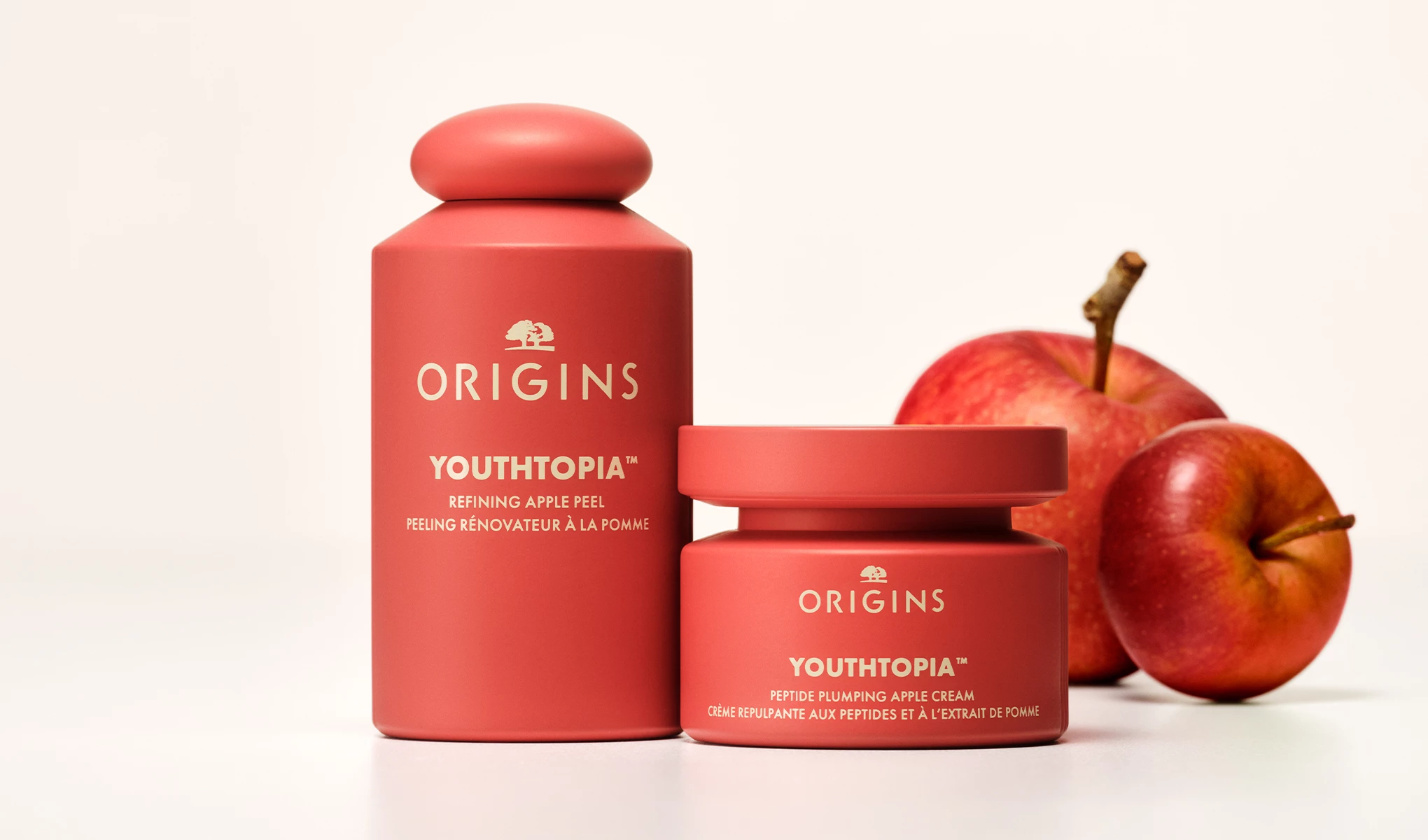 Origins Youthtopia Refining Apple Peel ja Peptide Plumping Apple Cream asetettuna punaisten omenoiden viereen vaaleaa taustaa vasten.