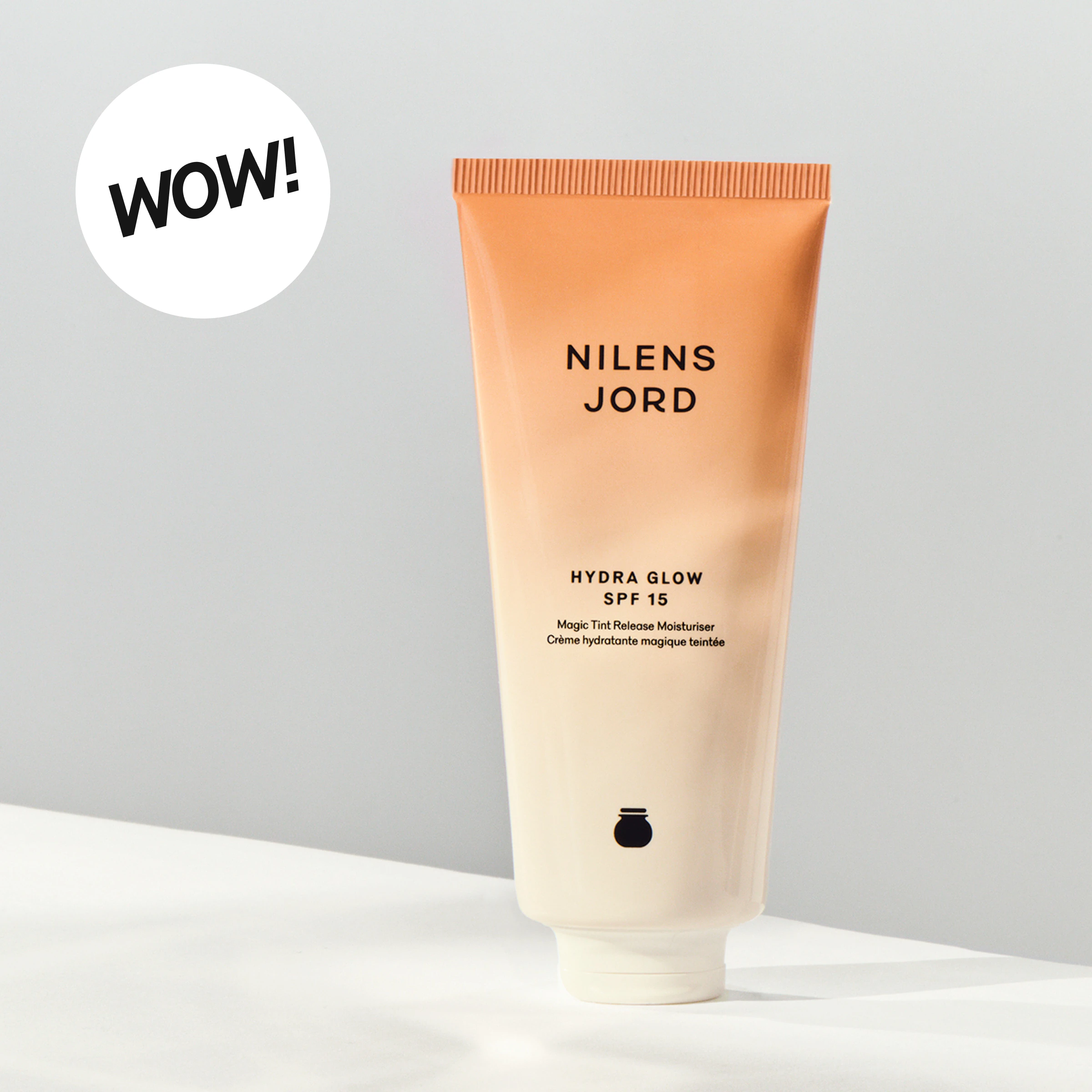 Tube fra Nilens Jord med teksten “NILENS JORD HYDRA GLOW SPF 15 Magic Tint Release Moisturiser Crème hydratante magique teintée”. En rund etikett med teksten “WOW!” vises ved siden av den fargede dagkremen.