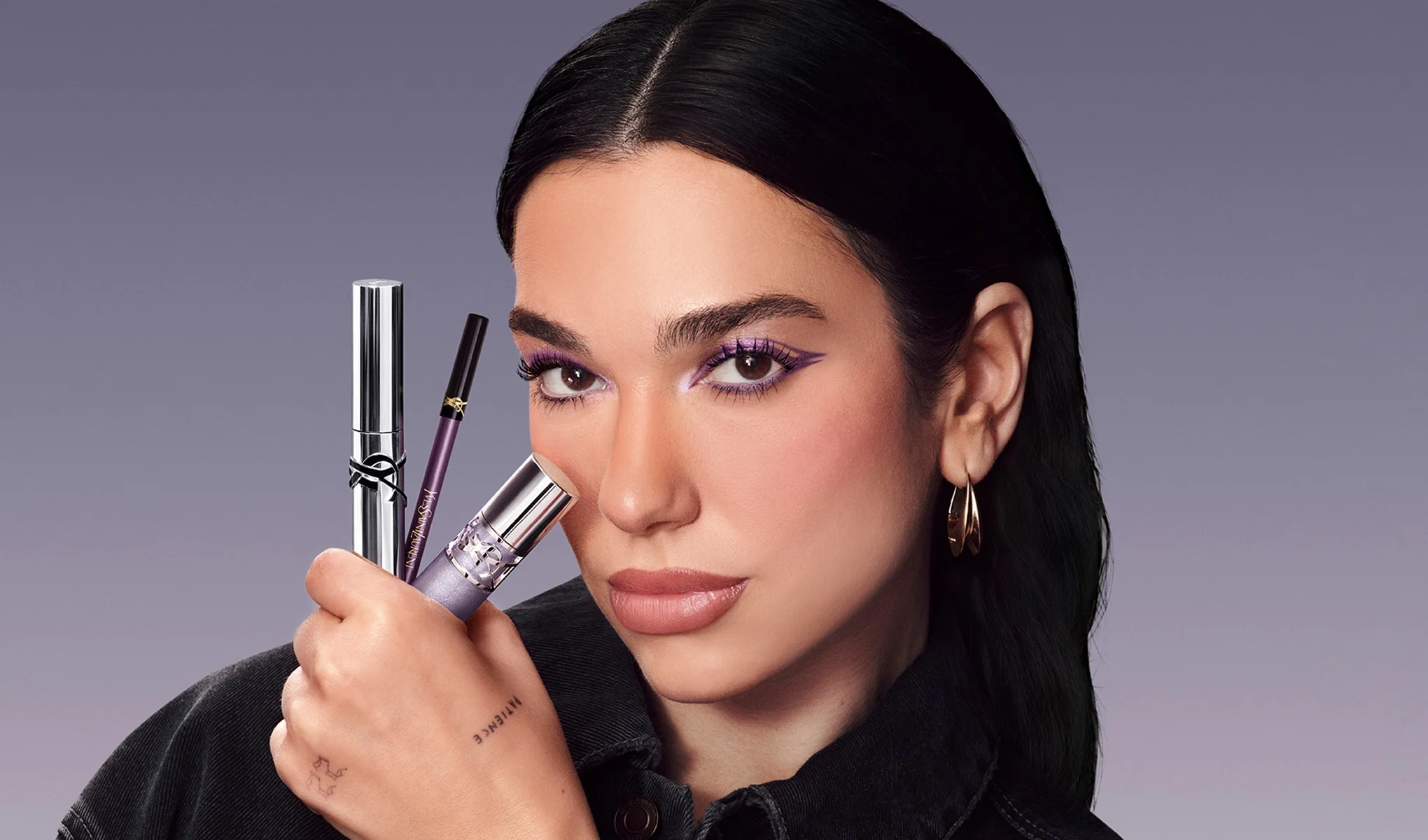 En person med lila ögonmakeup håller tre Yves Saint Laurent-produkter framför ansiktet: en silverfärgad mascara, en lila ögonpenna med YSL-logotyp och ett lila makeupstift. Personen bär guldfärgade örhängen och står mot en ljuslila bakgrund.