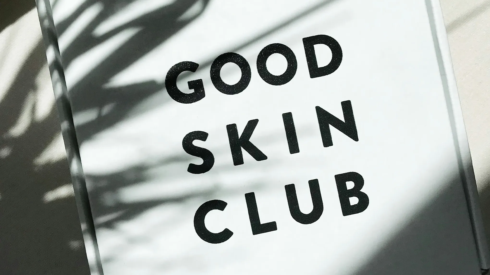 En vit skylt med texten "GOOD SKIN CLUB" i svarta bokstäver, delvis täckt av skuggor från växter.