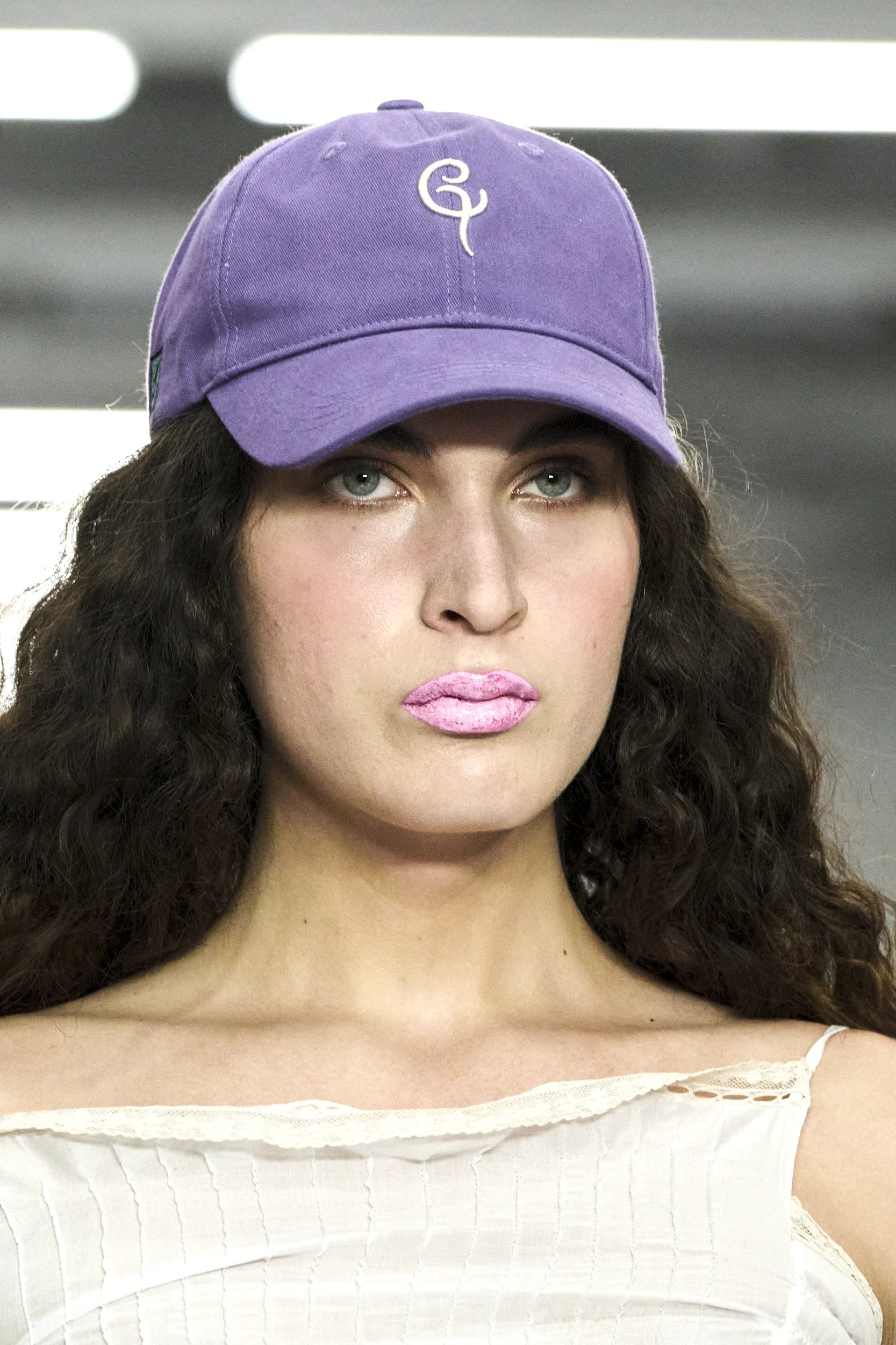 Nærbilde av en person med langt krøllete hår som bruker en lilla caps med hvit brodert logo og lys rosa leppestift. Personen har på en lys topp, med fokus på makeup som leppestift og ansikt foran en bakgrunn med lysrør.