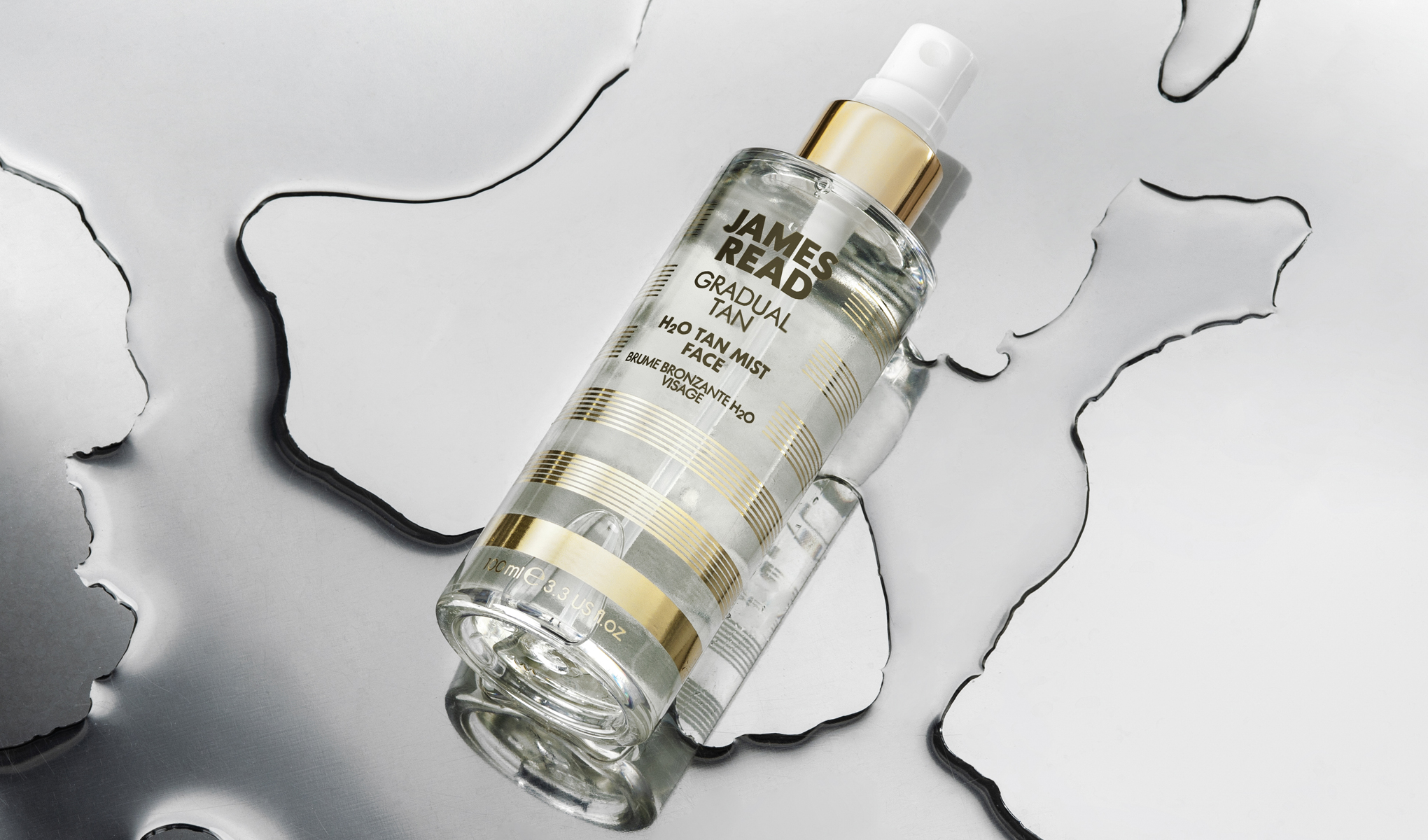 Pullo James Read Gradual Tan H2O Tan Mist -rusketussumutetta kasvoille asetettuna heijastavalle pinnalle, jonka ympärillä on vesiroiskeita. Pullon muotoilussa on kultaisia ja valkoisia raitoja ja valkoinen suihkupumppu.