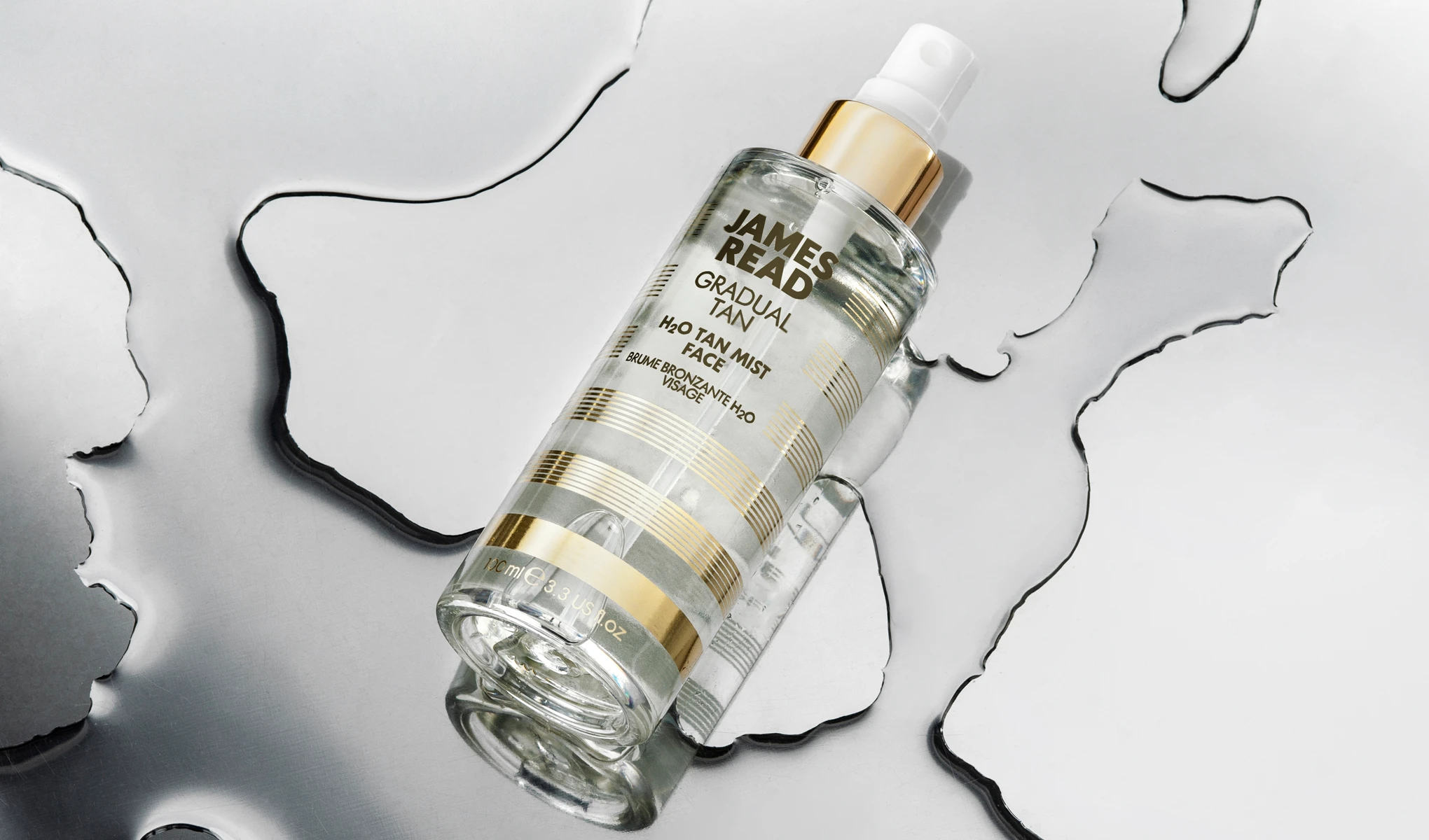 Pullo James Read Gradual Tan H2O Tan Mist -rusketussumutetta kasvoille asetettuna heijastavalle pinnalle, jonka ympärillä on vesiroiskeita. Pullon muotoilussa on kultaisia ja valkoisia raitoja ja valkoinen suihkupumppu.