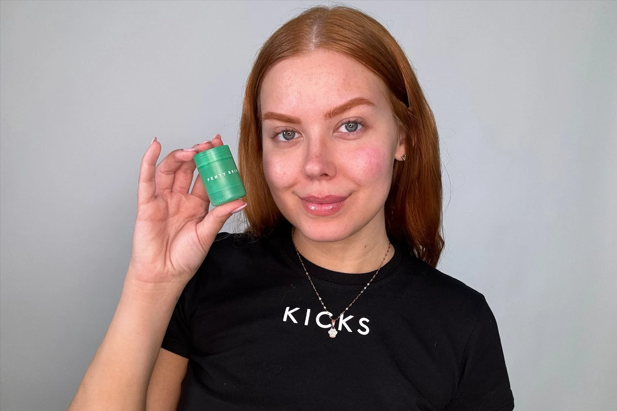 En närbild på en person med långt rött hår som håller i en grön burk med texten "FENTY SKIN" mot en grå bakgrund.