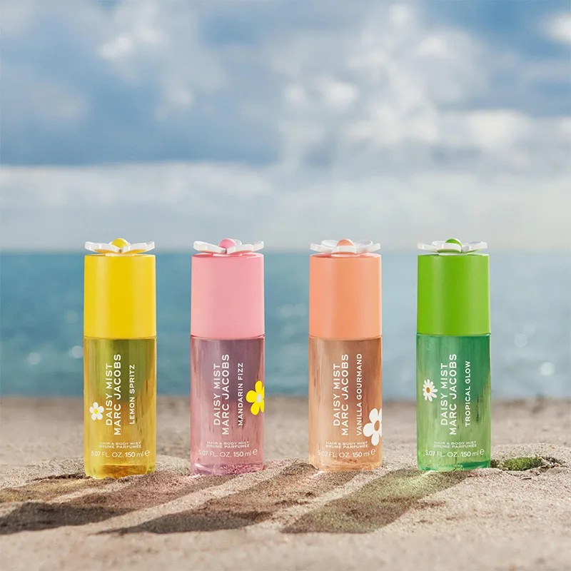 Neljä Marc Jacobs Daisy Mist Hair & Body Mist -tuotetta seisoo hiekalla meren edessä. Pullot ovat Lemon Spritz, Mandarin Fizz, Vanilla Gourmand ja Tropical Glow keltaisen, vaaleanpunaisen, persikan ja vihreän sävyissä, ja niissä on kukkamaiset korkit sekä näkyvä tuoteteksti.