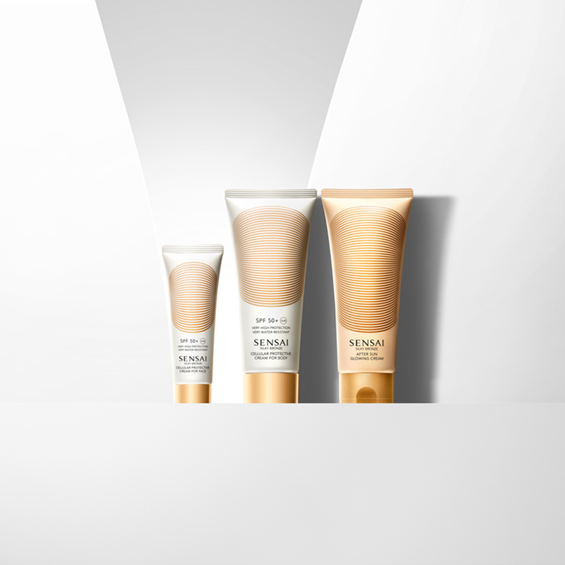 Tre tuber med SENSAIs hudvårdsprodukter står på en vit yta med en ljus och minimalistisk bakgrund. Produkterna har guldfärgade detaljer. Den vänstra tuben är mindre och vit, den mellersta tuben är medelstor och vit, och den högra tuben är större och guld. Bakgrunden har en triangulär ljuseffekt som framhäver produkterna.
