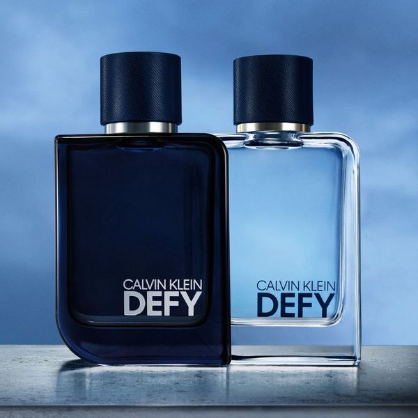 Två parfymer från Calvin Klein med namnet "Defy". Den ena flaskan är mörkblå och den andra är ljusblå, båda med mörkblå korkar. Flaskorna är placerade på en yta med en blå himmel i bakgrunden.