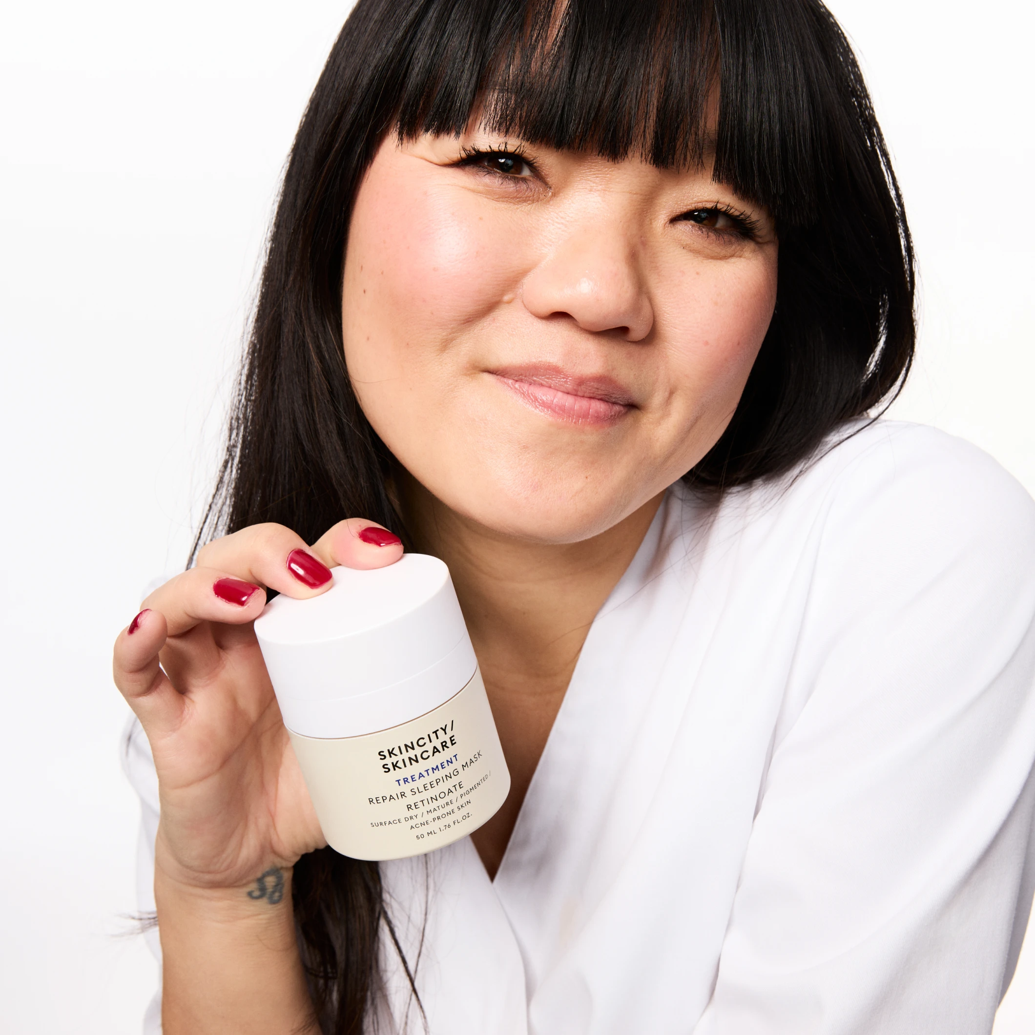 En person holder opp en krukke med Skincity Skincare Repair Retinol Sleeping Mask, kledd i hvit skjorte og med rødlakkerte negler.