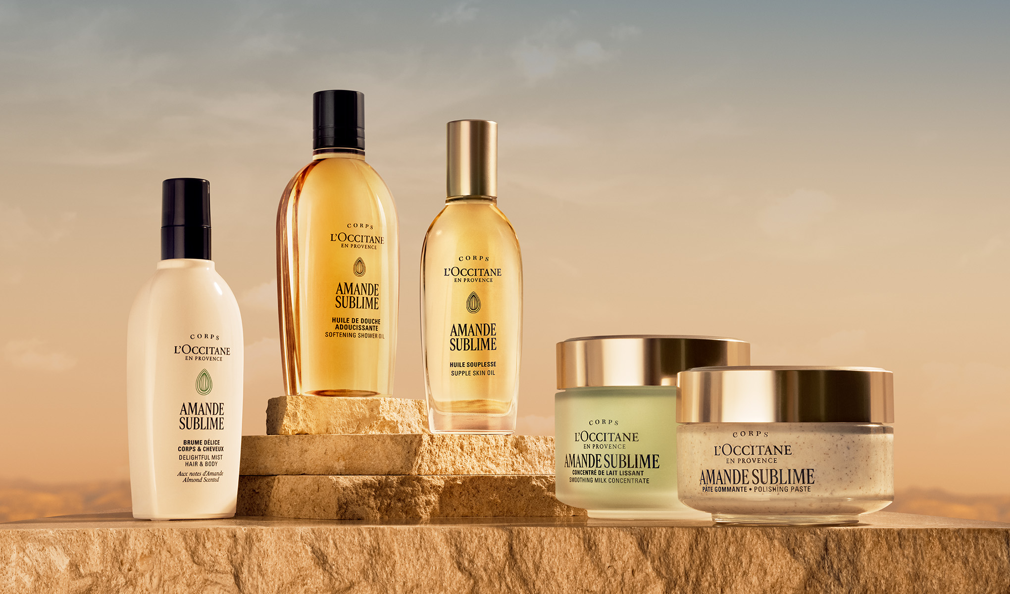 Flera kroppsvårdsprodukter från L’OCCITANE en Provence i serien “Amande Sublime” står på stenblock i varmt ljus, med text som “Huile de Douche Adoucissante Softening Shower Oil”, “Huile Souplesse Supple Skin Oil”, “Concentré de Lait Lissant Smoothing Milk Concentrate”, “Pâte Gommante Polishing Paste” och “Brume Délice Corps & Cheveux Delightful Mist Hair & Body”.