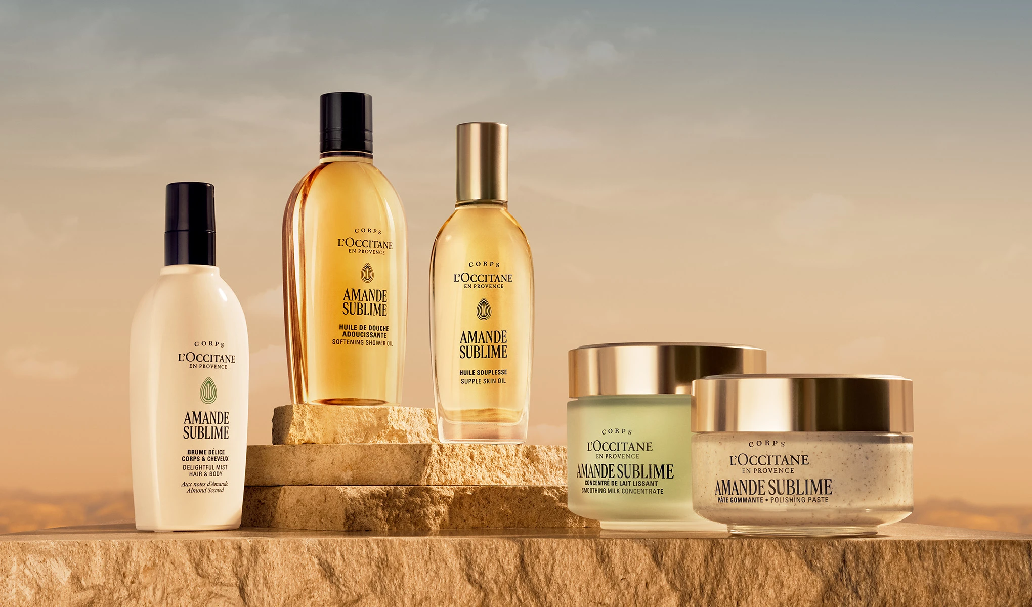 Useita L’OCCITANE en Provence -vartalonhoitotuotteita “Amande Sublime” -sarjasta on aseteltu kivitasoille lämpimässä valossa, teksteillä “Huile de Douche Adoucissante Softening Shower Oil”, “Huile Souplesse Supple Skin Oil”, “Concentré de Lait Lissant Smoothing Milk Concentrate”, “Pâte Gommante Polishing Paste” ja “Brume Délice Corps & Cheveux Delightful Mist Hair & Body”.