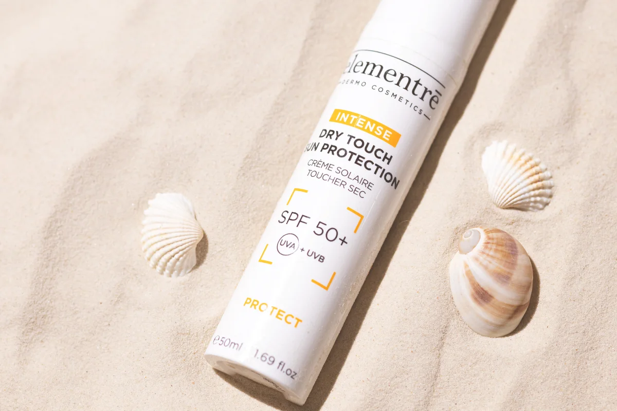 En flaske med intensiv solkrem med SPF 50+ ligger på sanden, omgitt av vakre skjell. 