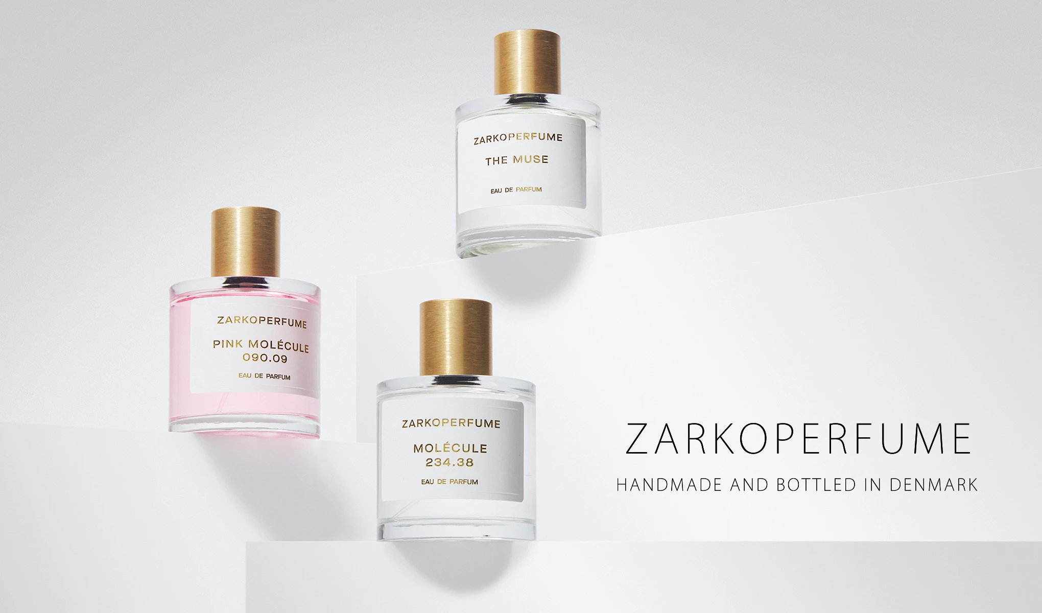 Tre ZARKOPERFUME Eau de Parfum-flasker står på hvite podier: Pink Molécule 090.09, Molécule 234.38 og The Muse. Synlig tekst: "ZARKOPERFUME", "PINK MOLÉCULE 090.09", "MOLÉCULE 234.38", "THE MUSE", "EAU DE PARFUM", "HANDMADE AND BOTTLED IN DENMARK" og "ZARKOPERFUME".