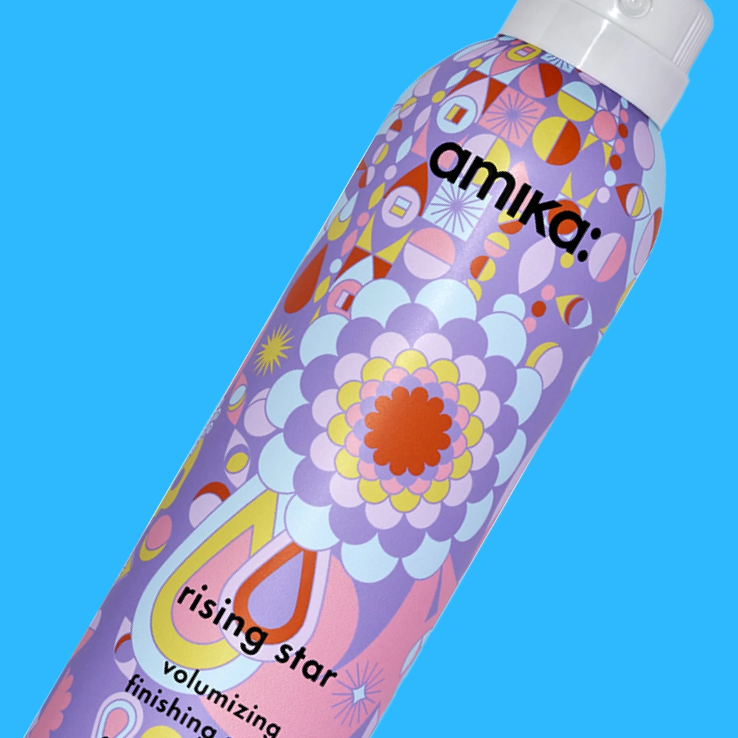 Amika Rising Star Volumizing Finishing Spray i lila sprayflaska med färgstarkt mönster mot blå bakgrund. På förpackningen syns texterna ”amika:”, ”rising star”, ”volumizing finishing spray”.