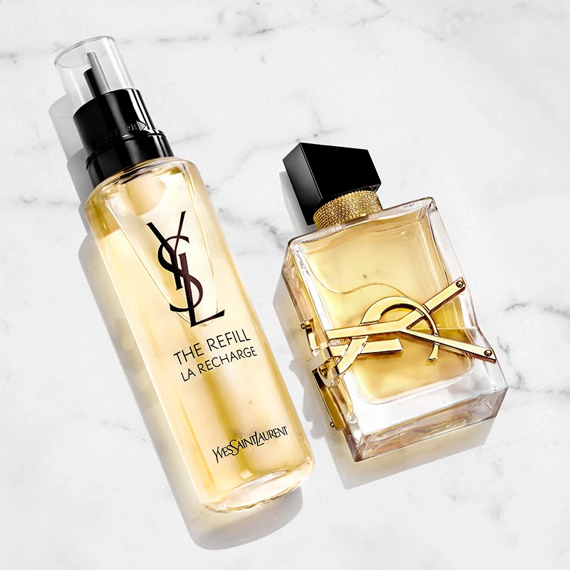 Kaksi Yves Saint Laurent -hajuvesipulloa valkoisella marmoritaustalla. Toinen pullo on täydennyspullo, jossa on YSL-logo mustalla. Toinen pullo on läpinäkyvä ja siinä on kullanväristä nestettä, ja siinä on YSL-logo kullanvärisenä ja musta korkki.