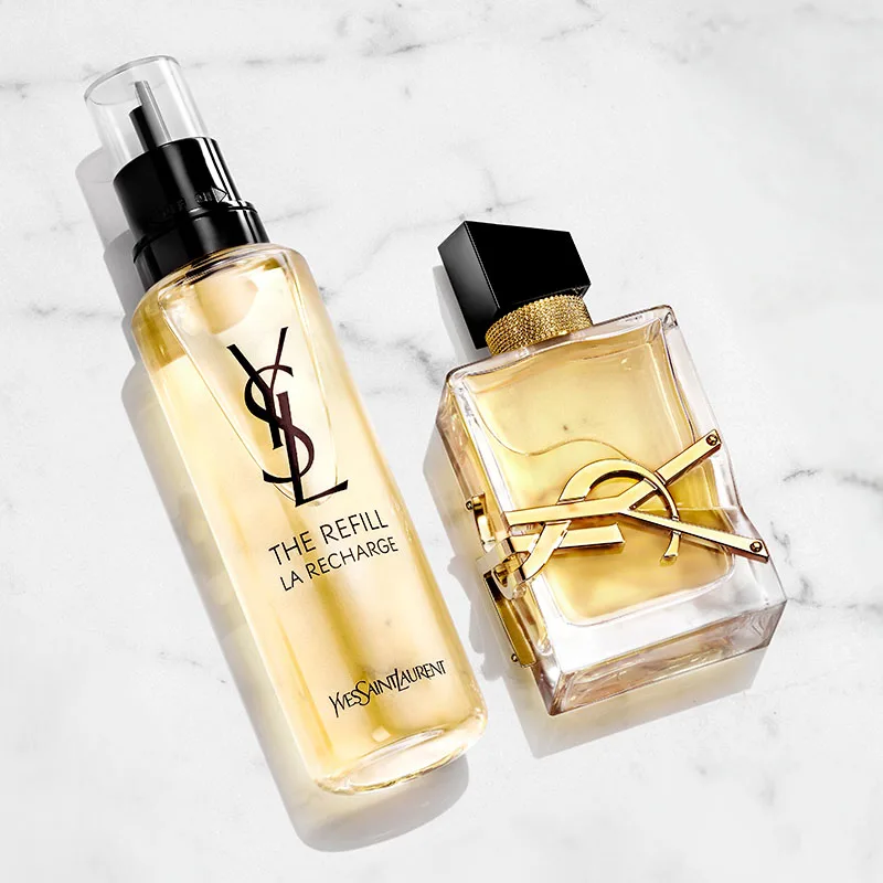 Två parfymflaskor från Yves Saint Laurent, liggande på en vit marmorbakgrund. Den ena flaskan är en refillflaska med YSL-logotypen i svart. Den andra flaskan är genomskinlig med en guldfärgad vätska och har YSL-logotypen i guld samt en svart kork.