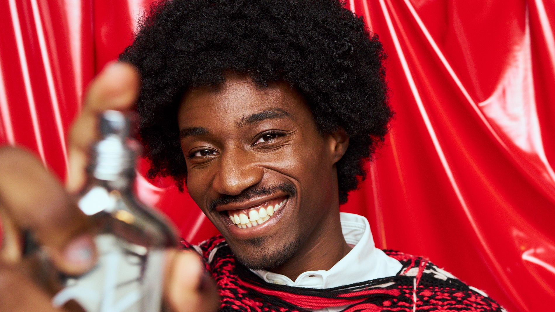 Kuva, joka näyttää henkilön, jolla on tummat kiharat hiukset ja joka pitää hajuvettä. Henkilöllä on valkoinen paita ja punainen ja musta neule. Tausta on punainen.