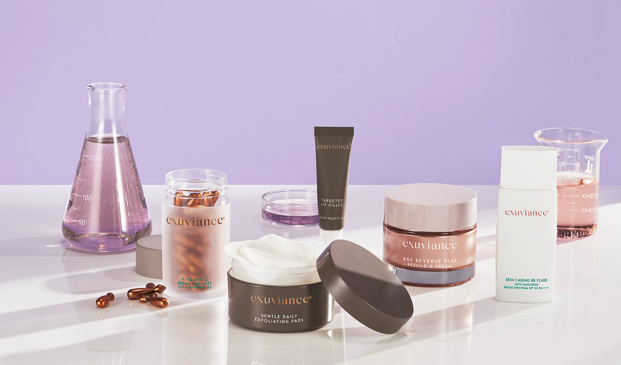 En samling hudpleieprodukter fra Exuviance er utstilt på et hvitt bord foran en lyslilla bakgrunn. Bak produktene står laboratorieglass med rosa væske.