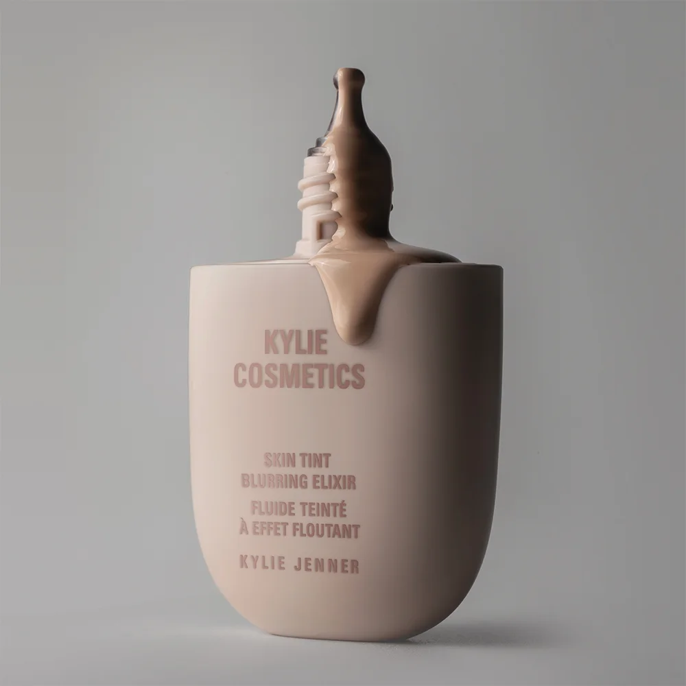 En flaska från Kylie Cosmetics med texten "Skin Tint Blurring Elixir" står mot en ljus bakgrund. Vätska rinner ut från flaskans öppning, vilket skapar ett elegant och inbjudande utseende.