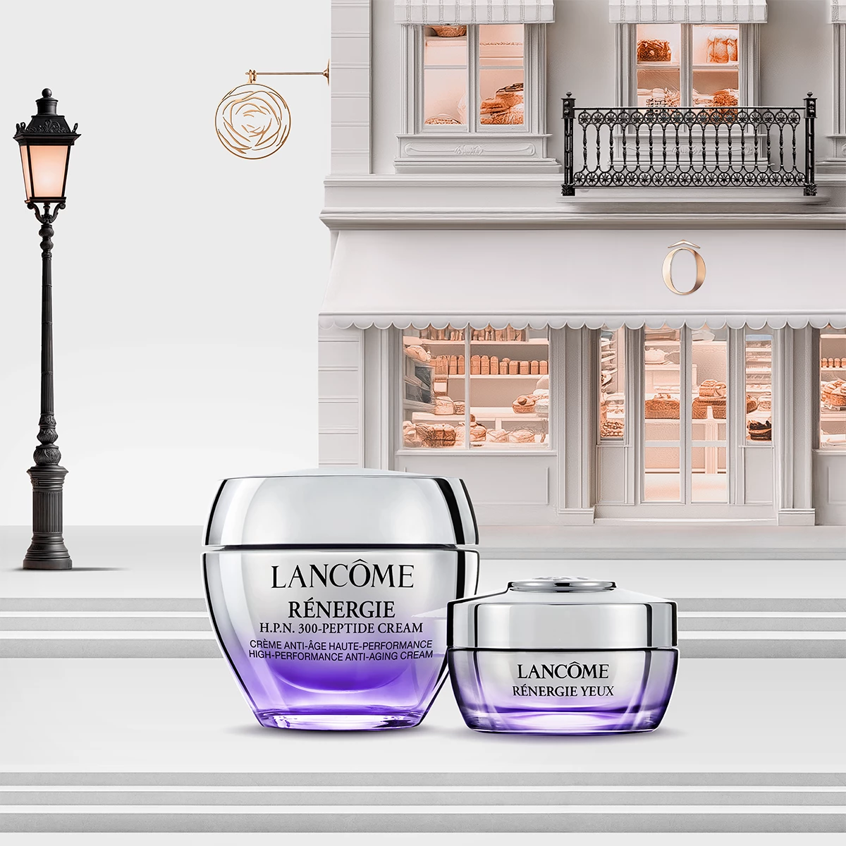 Kaksi Lancôme Rénergie ihonhoitotuotetta, joissa on violetti ja hopeanvärinen pakkaus, elegantin parvekkeella ja myymälän ikkunoilla varustetun rakennuksen edessä. Vasemmalla on lyhtypylväs.