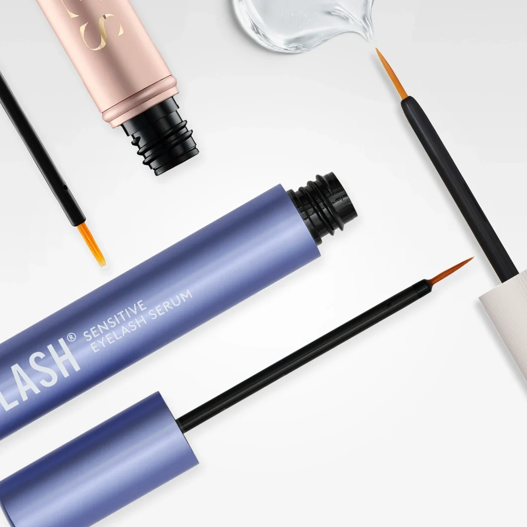 Øyevippeserum i tuber med teksten “LASH Sensitive Eyelash Serum” vises sammen med applikatorbørster og en dråpe transparent serum.
