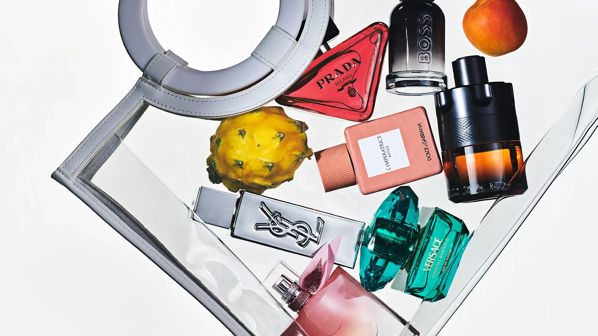 Flere parfymeflasker arrangert i en gjennomsiktig håndveske sammen med frukt. Synlige parfymer fra Prada, BOSS, Yves Saint Laurent, Dolce & Gabbana, Versace og Lancôme i ulike fargede glassflasker.