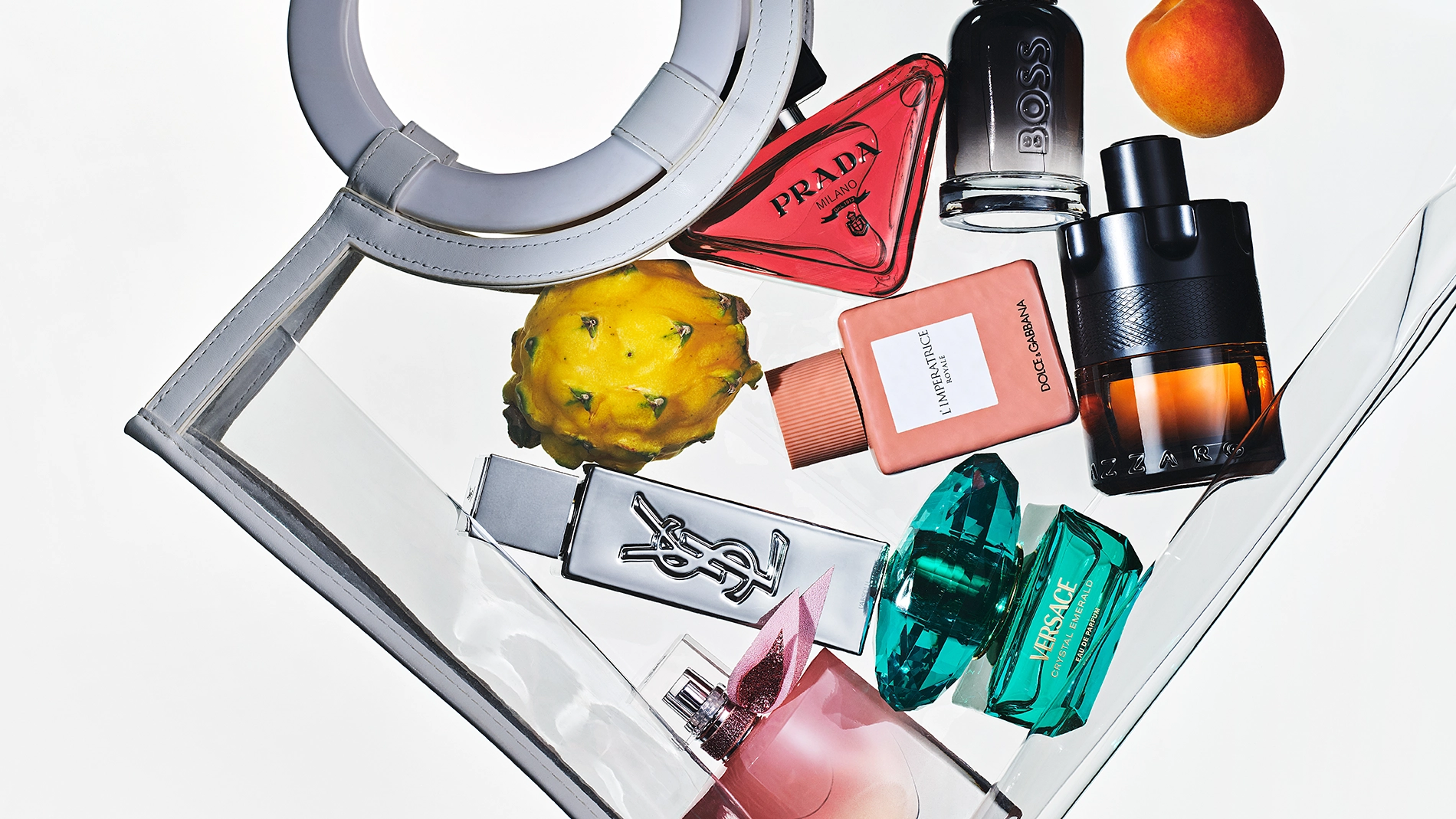 Flera parfymflaskor arrangerade i en transparent handväska tillsammans med frukt. Synliga parfymflaskor från Prada, BOSS, Yves Saint Laurent, Dolce & Gabbana, Versace och Lancôme med olika färgade glasflaskor.