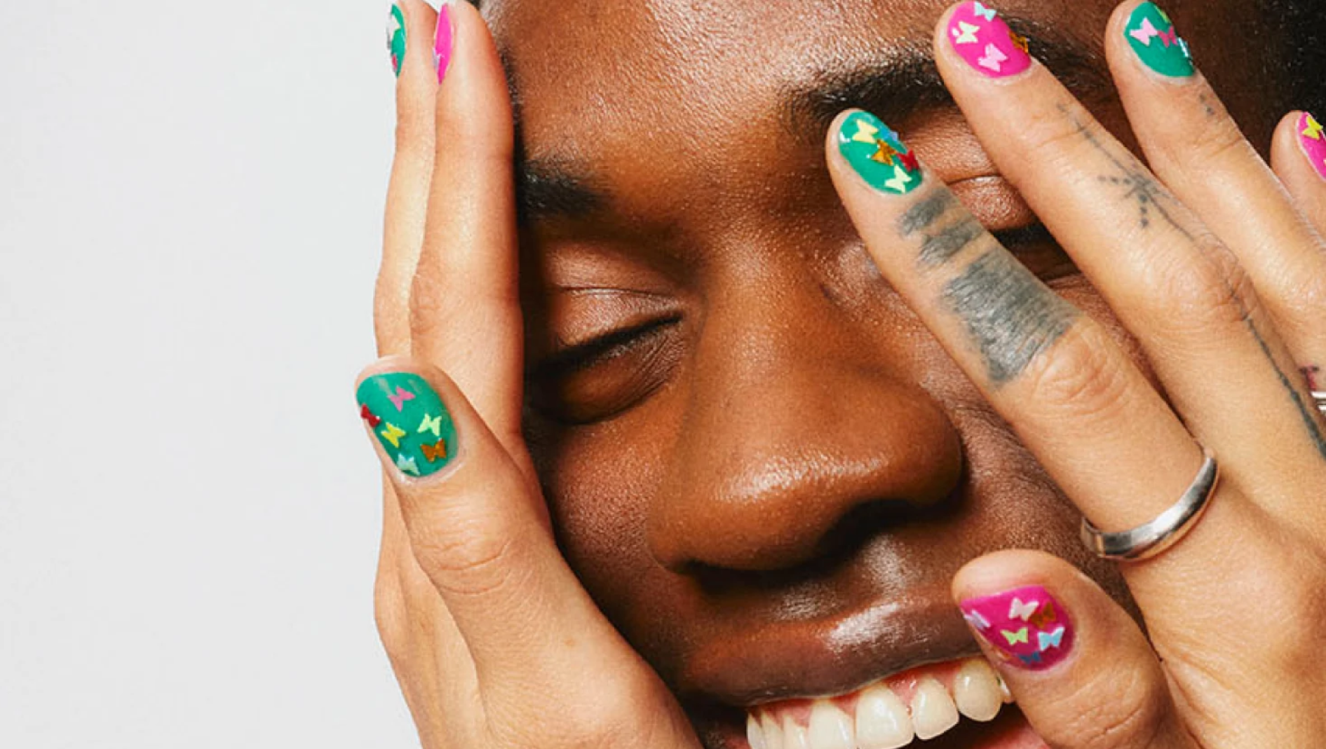 Lähikuva henkilöstä, jonka kädet ovat kasvoja vasten ja silmät suljettuina. Kynsissä on värikäs pinkki ja turkoosi nail art pienillä erivärisillä perhosilla, ja sormissa näkyy hopeanvärisiä sormuksia.