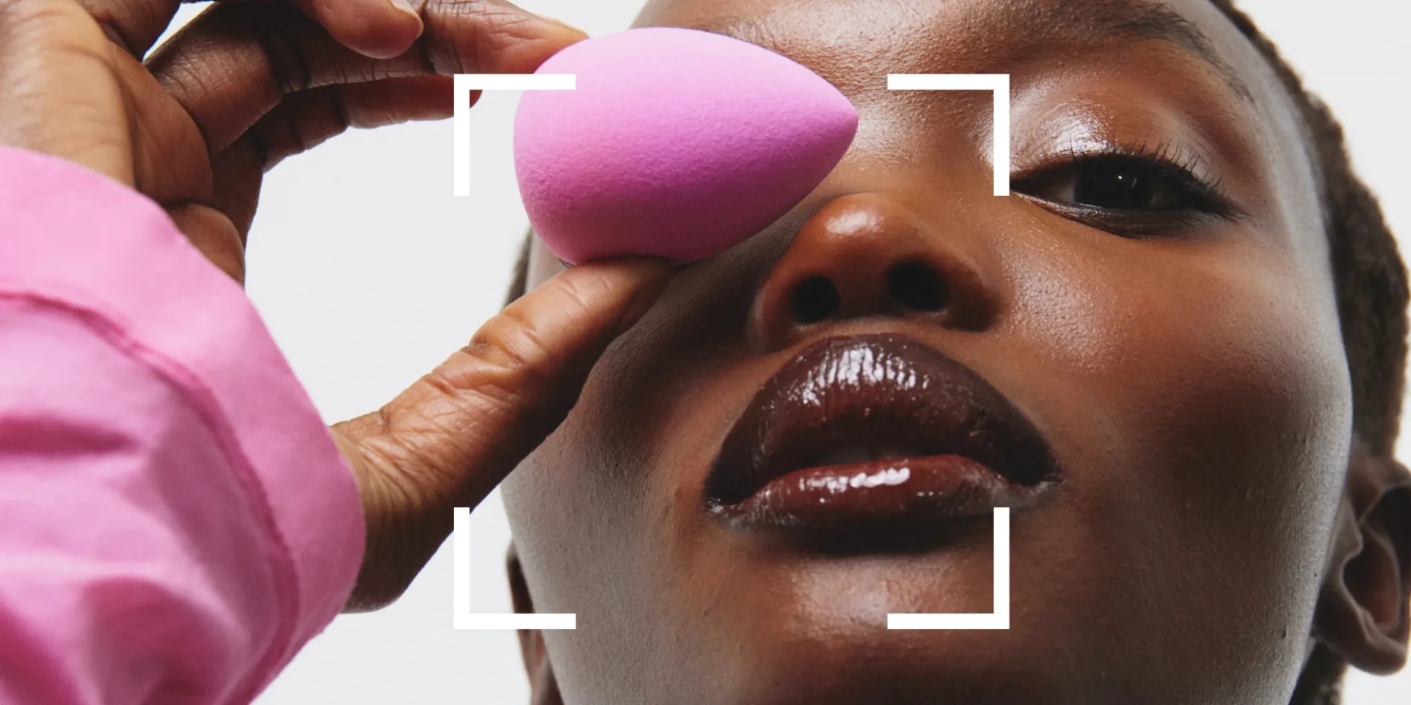 Närbild underifrån av en person som applicerar makeup med en rosa makeupsvamp mot ansiktet. Personen bär en rosa långärmad topp med hög krage. Vita markeringsramar syns runt ansiktet mot ljus bakgrund.
