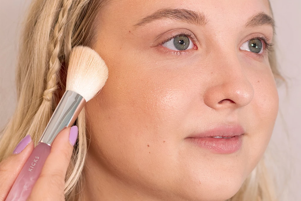 Blond kvinne med naturlig sminkebase, påfører highlighter på kinnet.