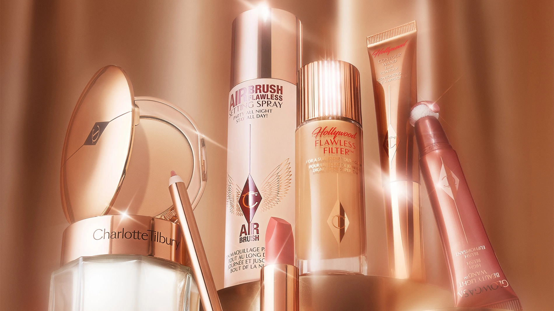 En samling av Charlotte Tilbury skjønnhetsprodukter, inkludert Airbrush Flawless Setting Spray, Hollywood Flawless Filter, Hollywood Contour Wand, Glowgasm Beauty Light Wand, en leppeblyant og en pudderkompakt. Produktene er elegant plassert og belyst med et varmt lys.