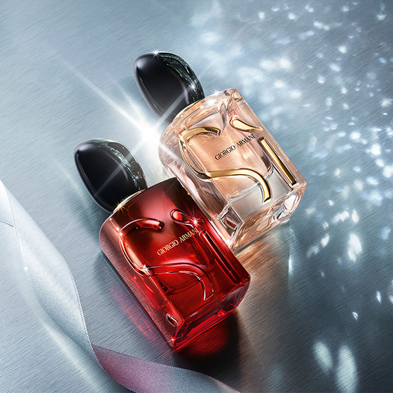 Två Giorgio Armani-parfymflaskor, en röd och en rosa, placerade på en silverfärgad yta. Bakgrunden har blåaktig belysning och ett silverfärgat band.