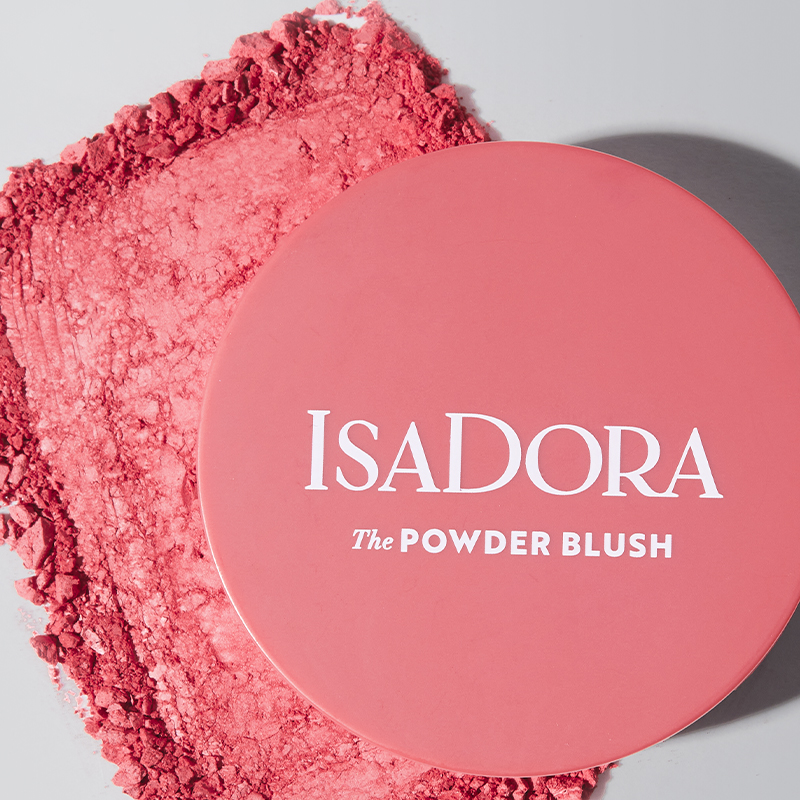 Pyöreä pakkaus, jossa on teksti ”IsaDora The Powder Blush” valkoisella. Pakkaus makaa pinnalla, jolle on levitetty vaaleanpunaista puuteria. Tausta on vaaleanharmaa.