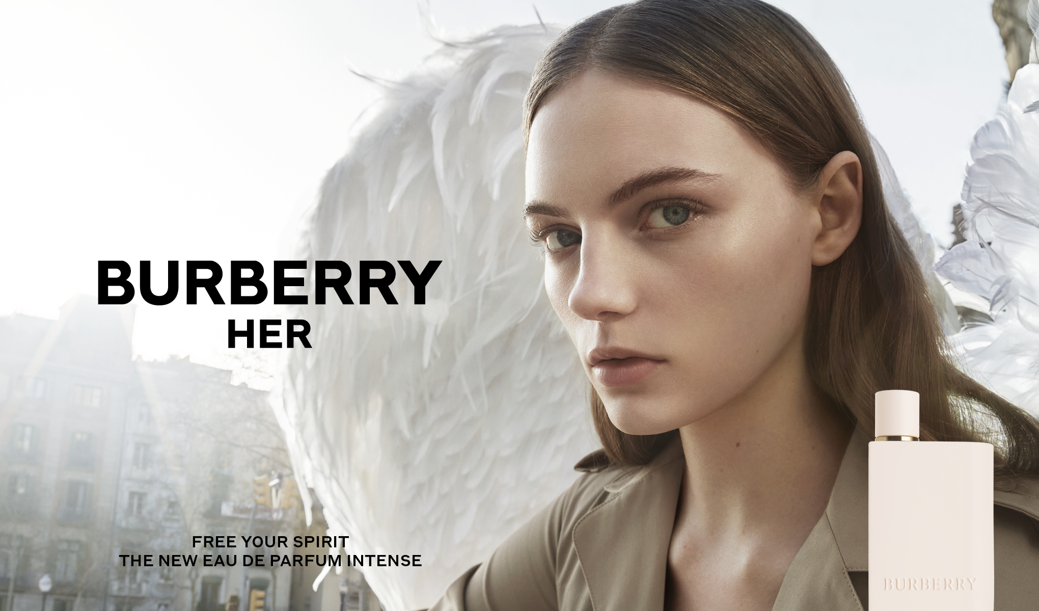 Ulkona seisoo nainen, jonka takana on suuria valkoisia höyheniä. Kuvan vasemmalla puolella on teksti ”Burberry Her”. Alareunassa on teksti ”Free Your Spirit The New Eau De Parfum Intense”. Vaaleanpunainen hajuvesipullo, jossa on teksti ”Burberry”, on sijoitettu oikeaan alakulmaan. Taustalla näkyy kaupunkiympäristö.