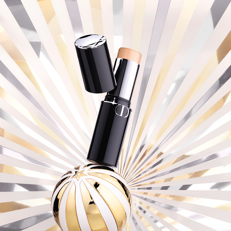 Dior foundation stick, med en bakgrunn av strålende linjer i gull, rosa og hvitt, samt en dekorativ gull- og hvit kule.