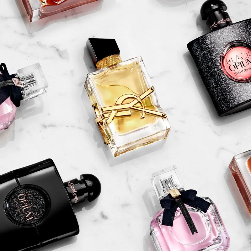 Fera parfymflaskor från Yves Saint Laurent. Flaskorna är elegant arrangerade på en vit marmorbakgrund. Den centrala flaskan är genomskinlig med en guldfärgad vätska och har YSL-logotypen i guld. Runt den finns andra flaskor i olika färger och former, inklusive svart, rosa och orange.